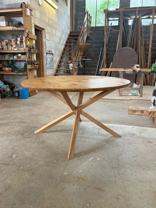 Table ronde en bois tressé dans un atelier, avec des étagères rustiques et des outils à l’arrière-plan.