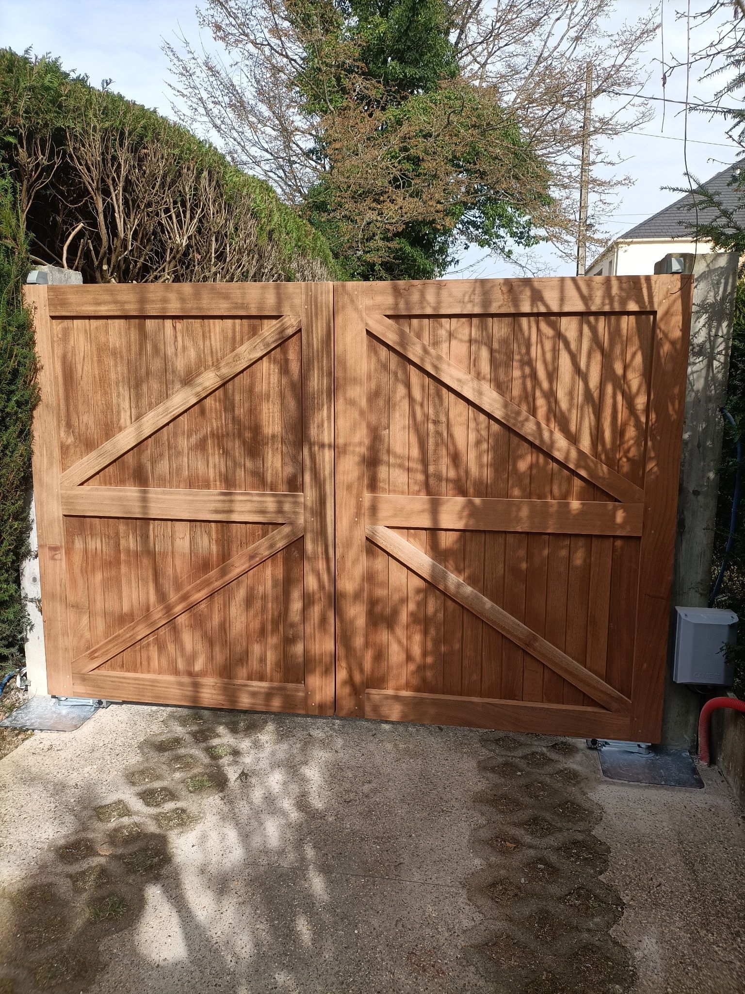 Portail double en bois avec des renforts diagonaux, installé entre des haies et une allée ensoleillée.