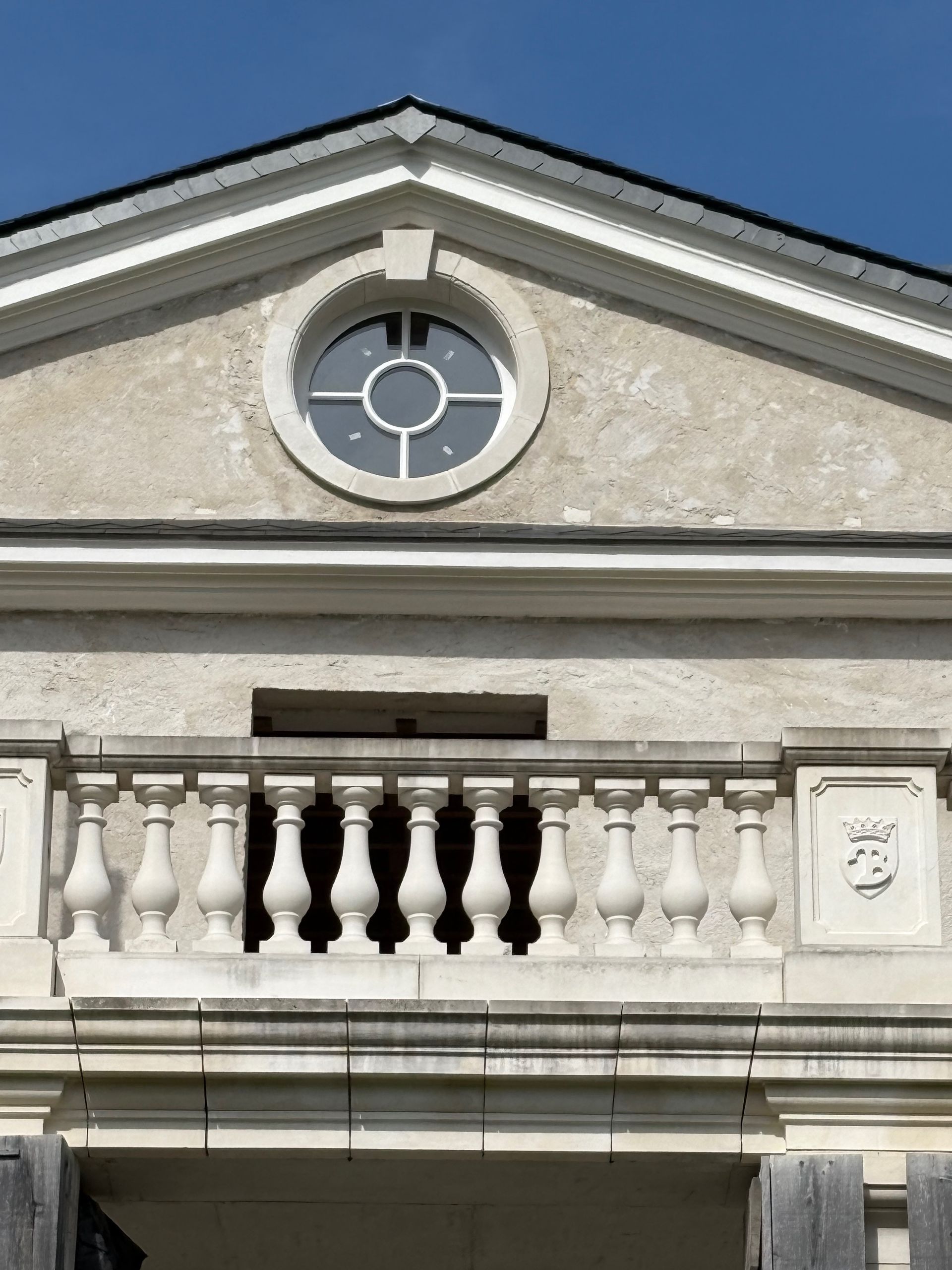 Gros plan sur la façade d’un bâtiment avec une fenêtre ronde, une rambarde de balcon blanche et un ciel bleu au-dessus.