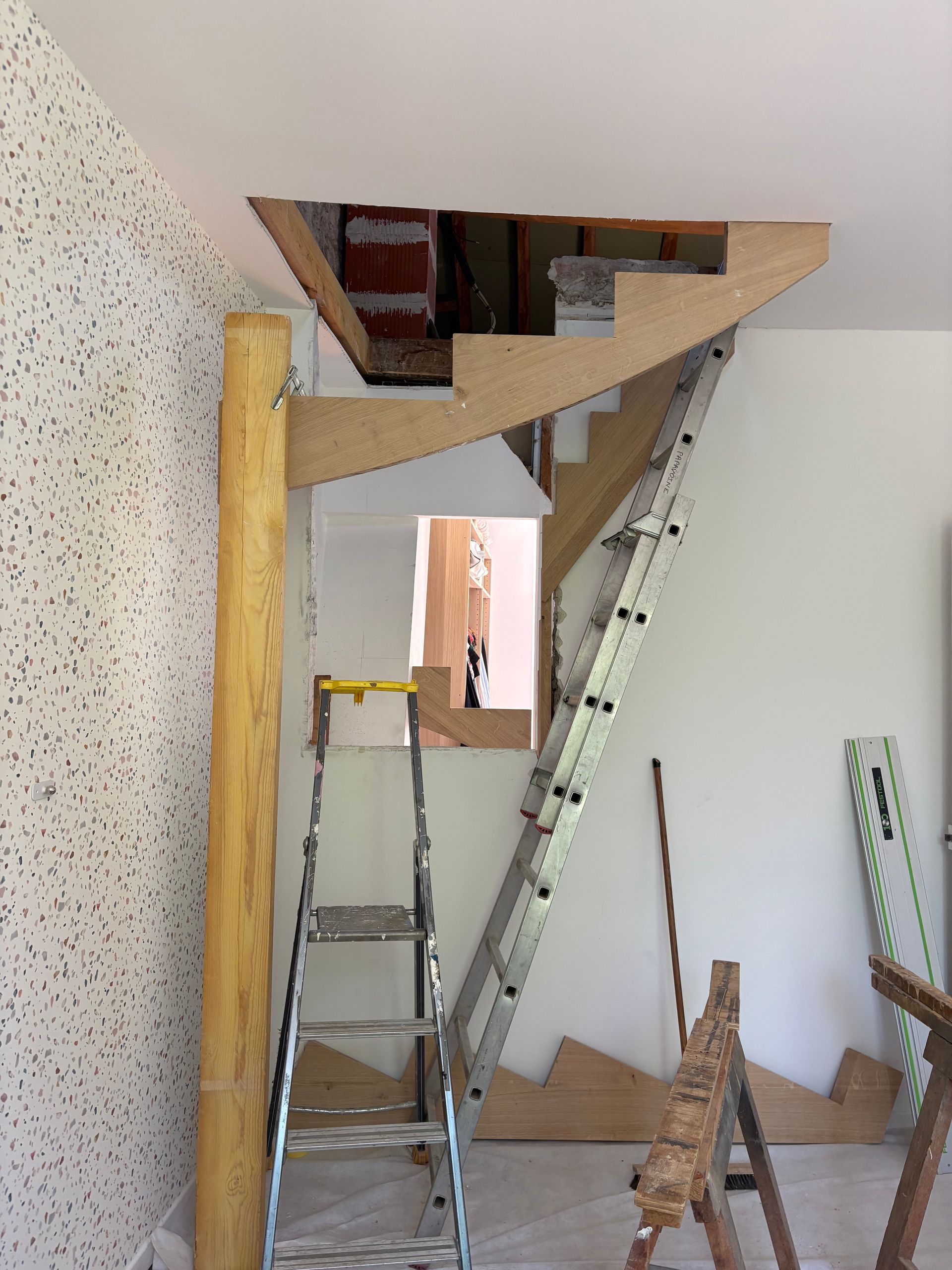 Cage d’escalier intérieure en cours de construction, avec une échelle, des marches inachevées et une charpente apparente.