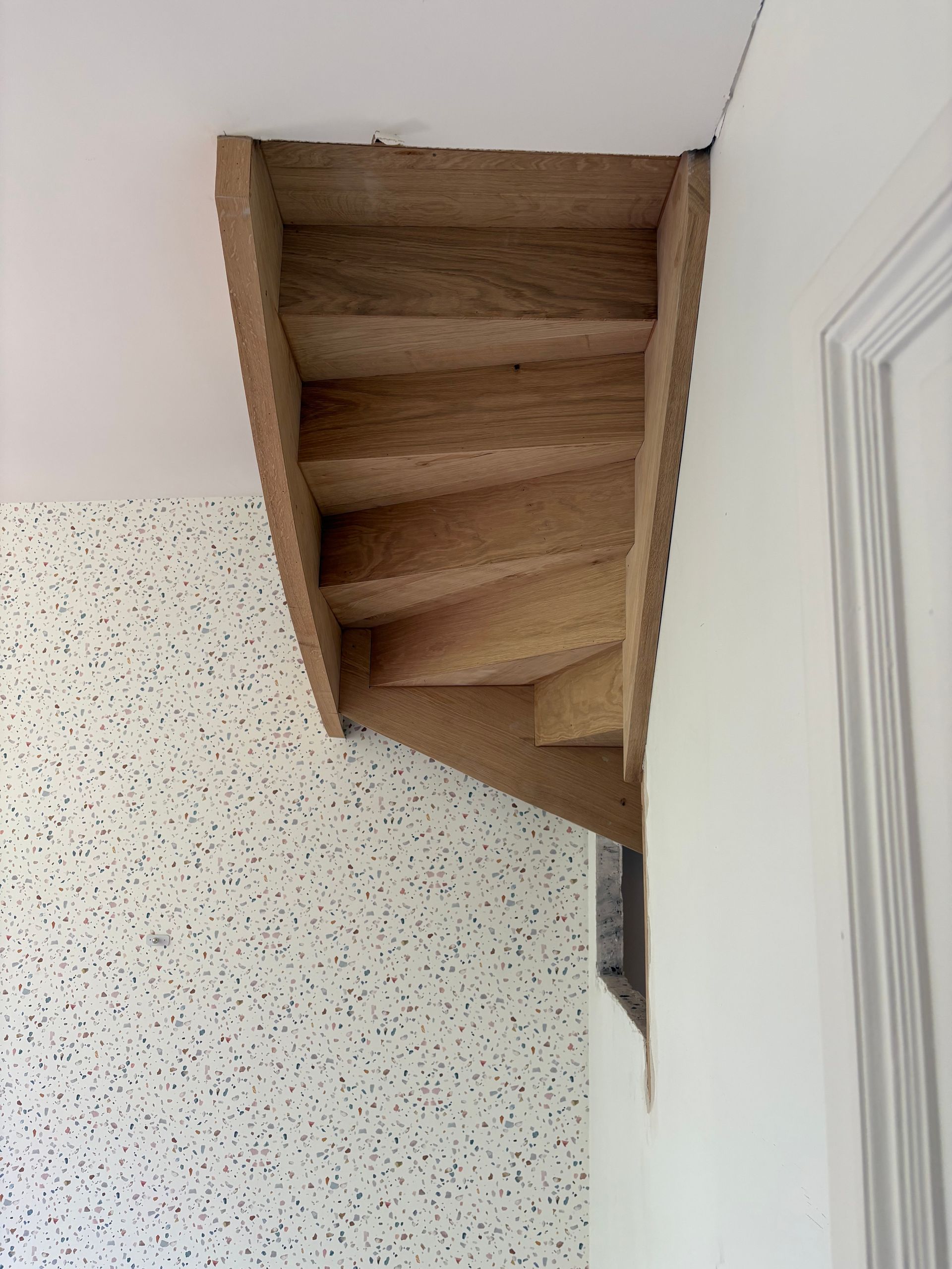 Étroite volée d’escalier en bois menant aux combles, qui monte en tournant dans un angle au-dessus d’un mur blanc recouvert de papier peint moucheté.