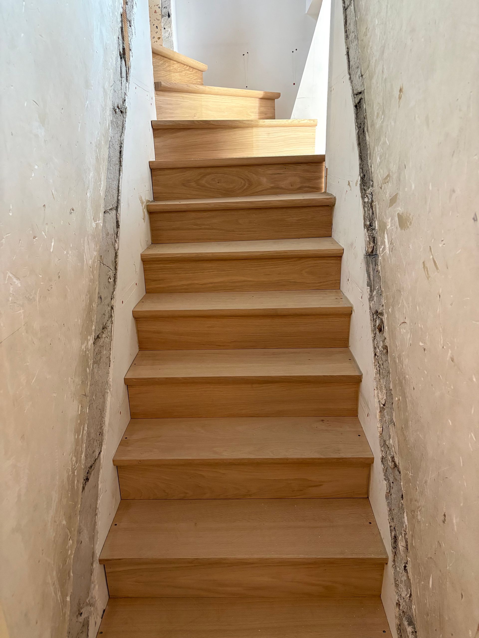 Escalier étroit en bois, murs blancs, dans un petit couloir intérieur.