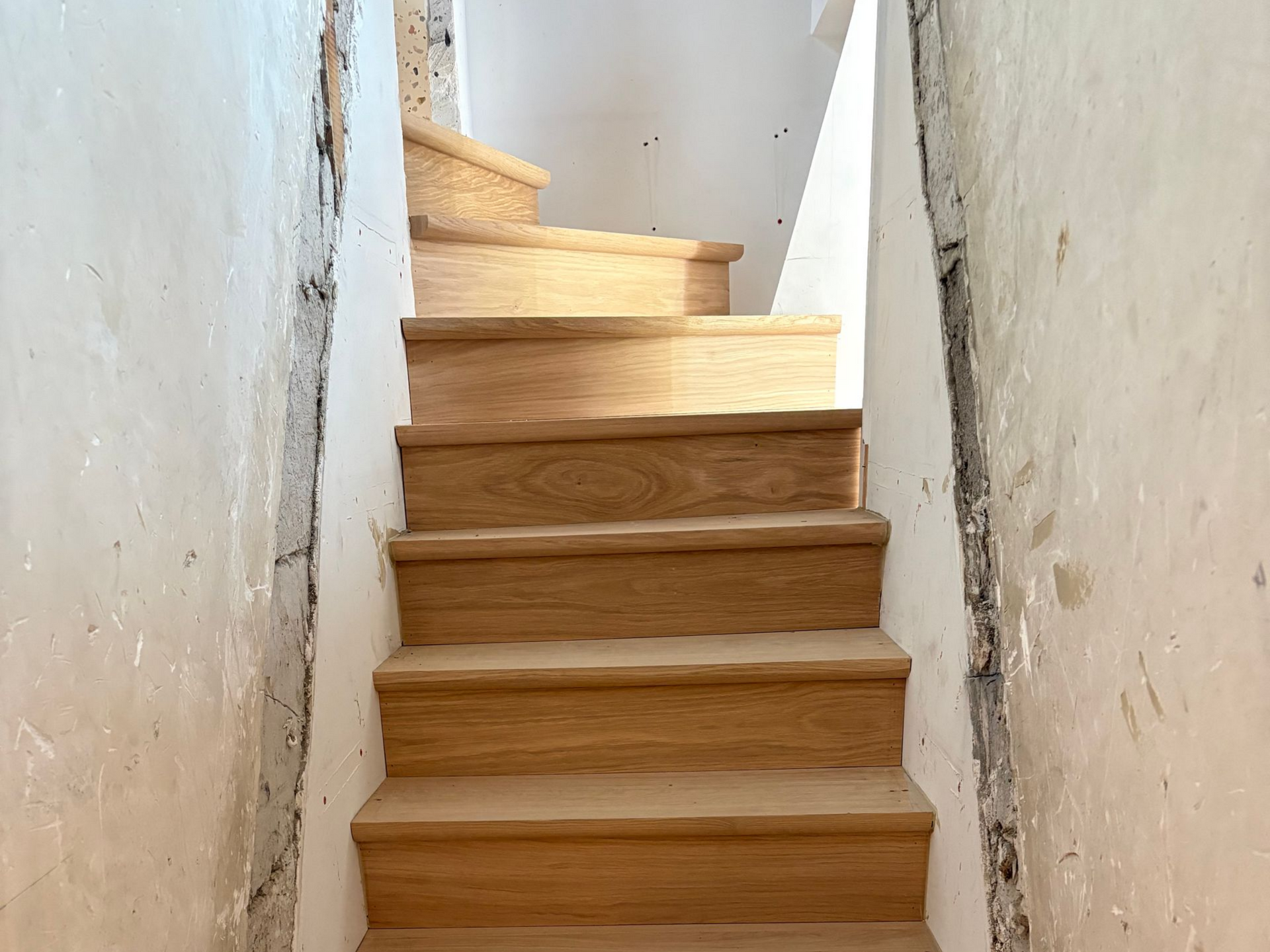 Escalier étroit en bois aux murs blancs, vu d’en bas.