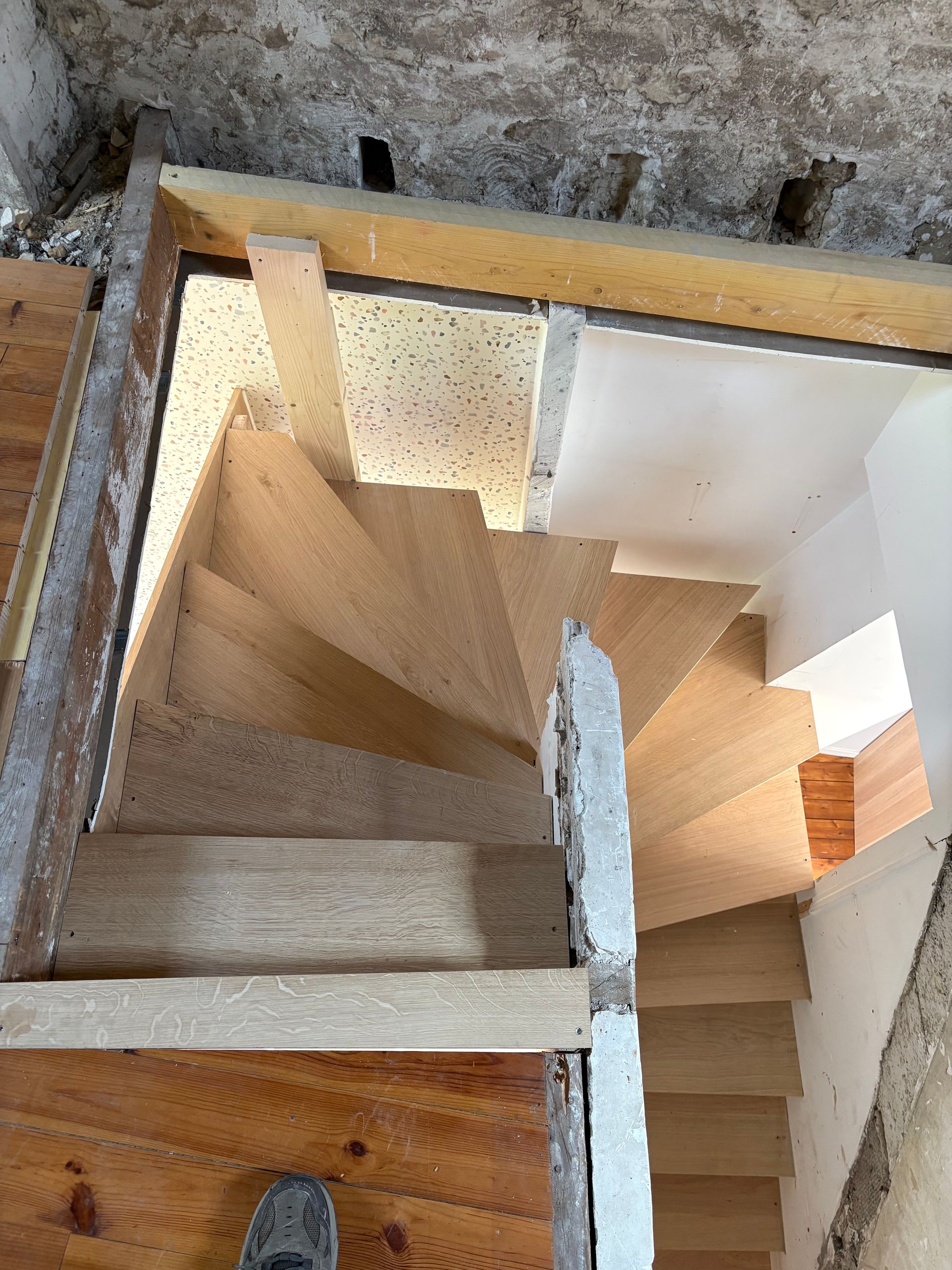 Vue plongeante sur un étroit escalier en colimaçon en bois, avec des murs blancs et un palier en béton, vu à travers une ouverture.