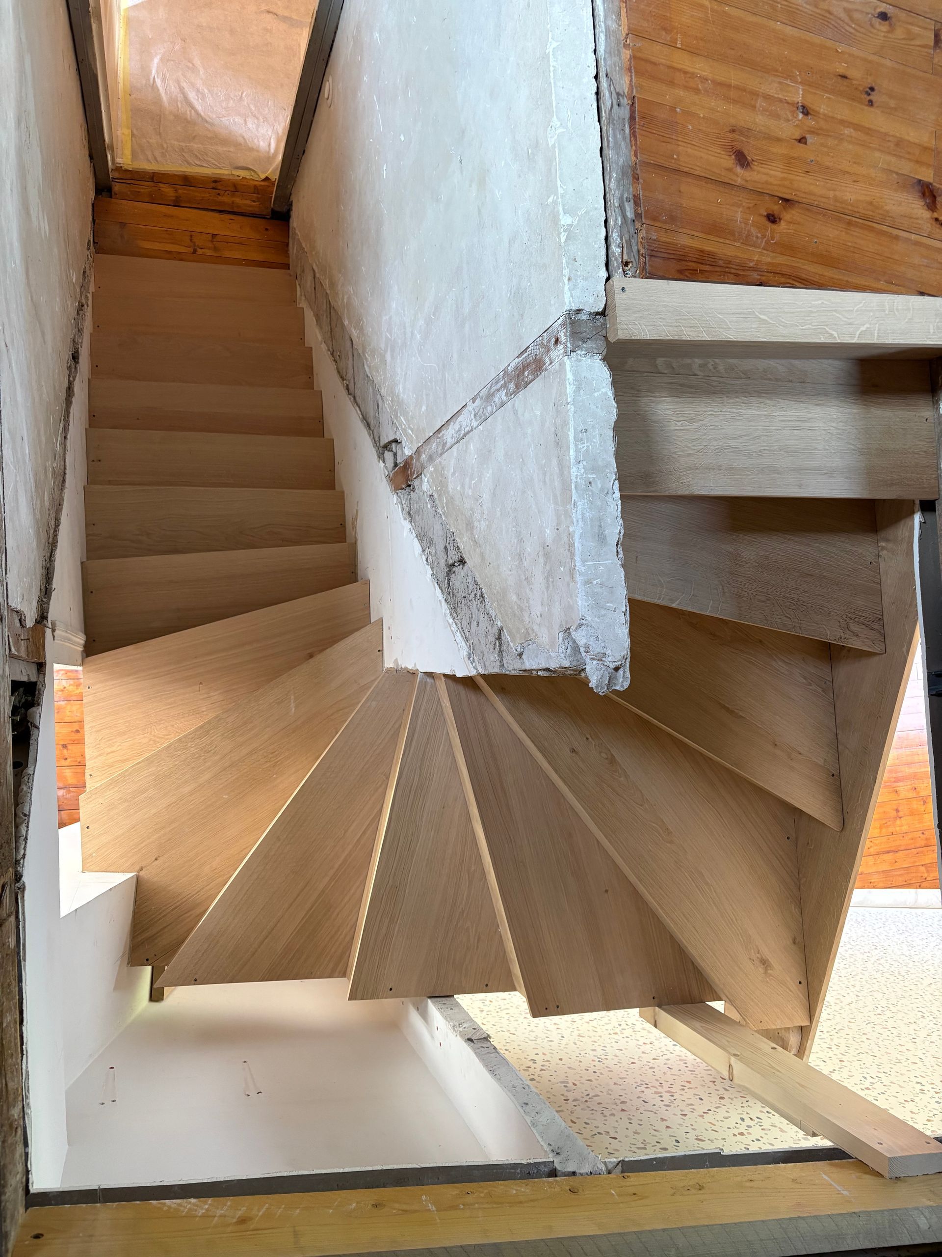 Vue plongeante d’un étroit escalier en colimaçon, avec des murs blancs et des marches en bois.