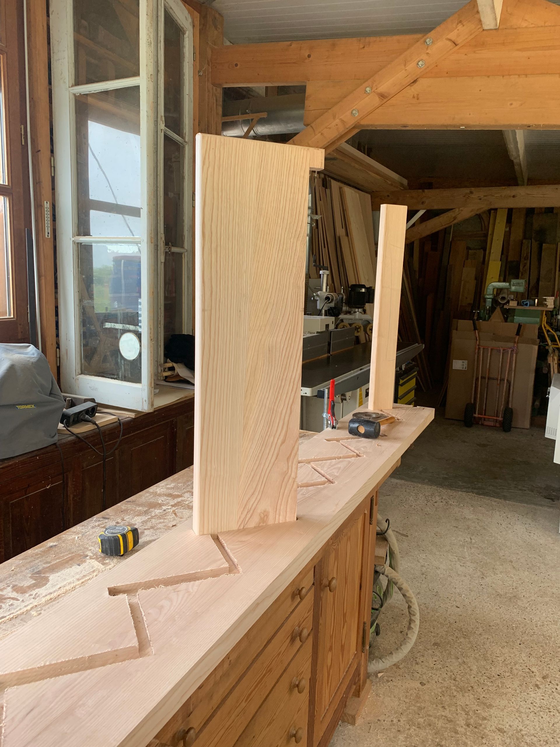 Armoire en bois dans un atelier, avec des outils sur un établi et une ossature en bois brut à proximité.