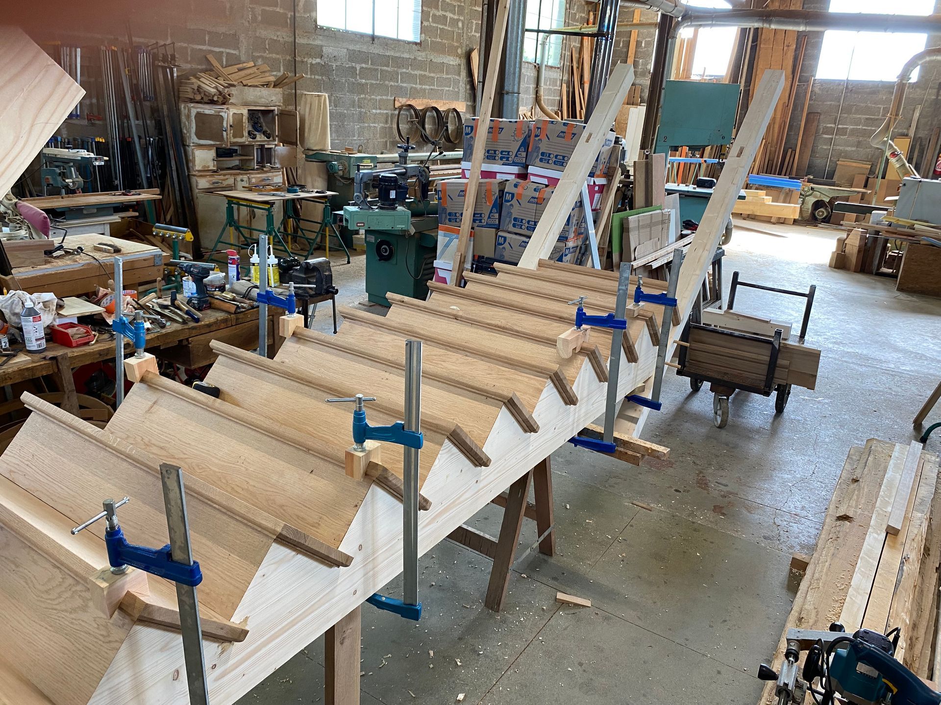 Atelier de menuiserie avec des rangées de pièces en bois non finies serrées par des serre-joints sur un long établi.