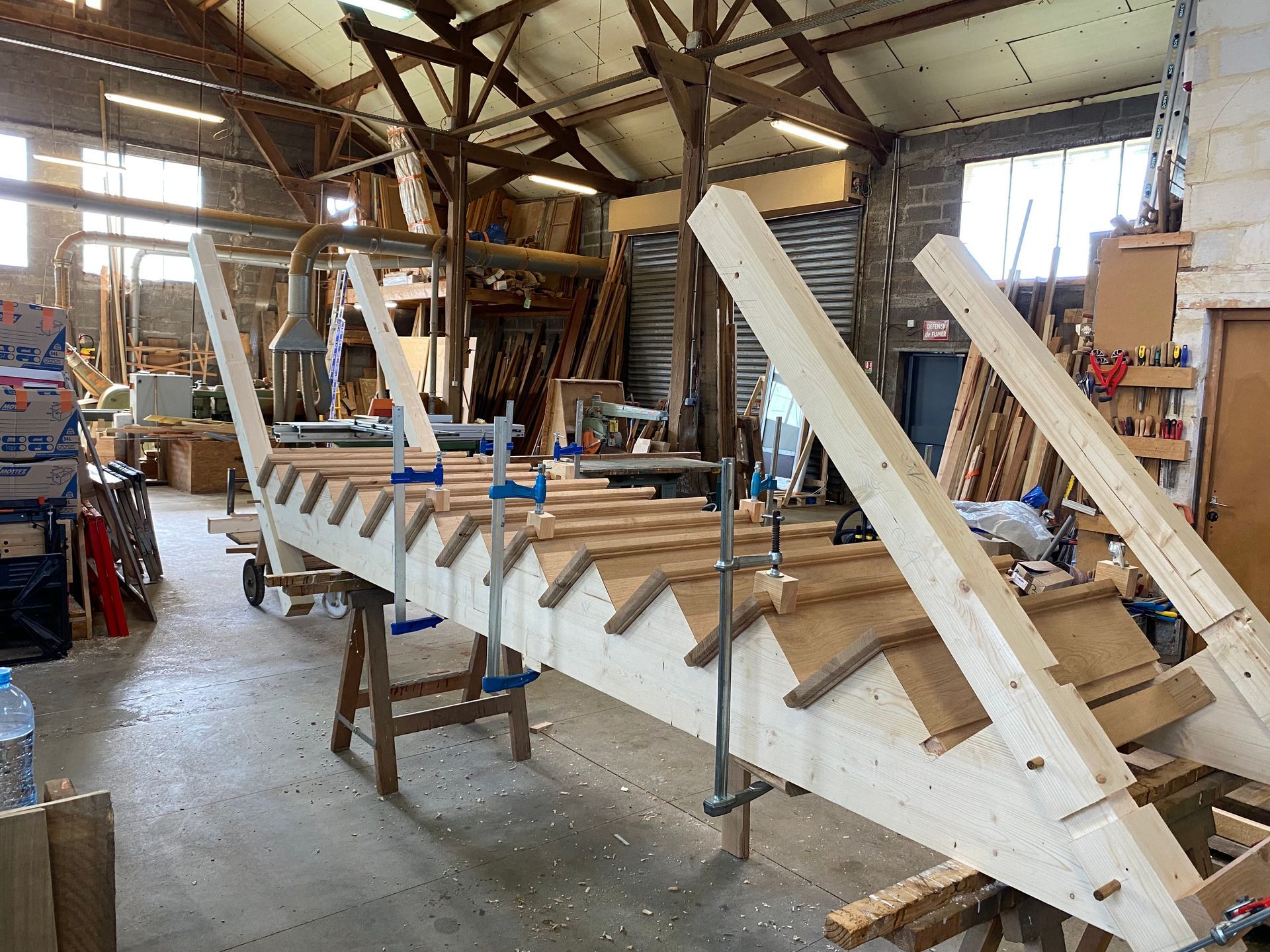 Atelier avec un bateau en bois en cours de construction, maintenu par des serre-joints dans un hangar industriel encombré.