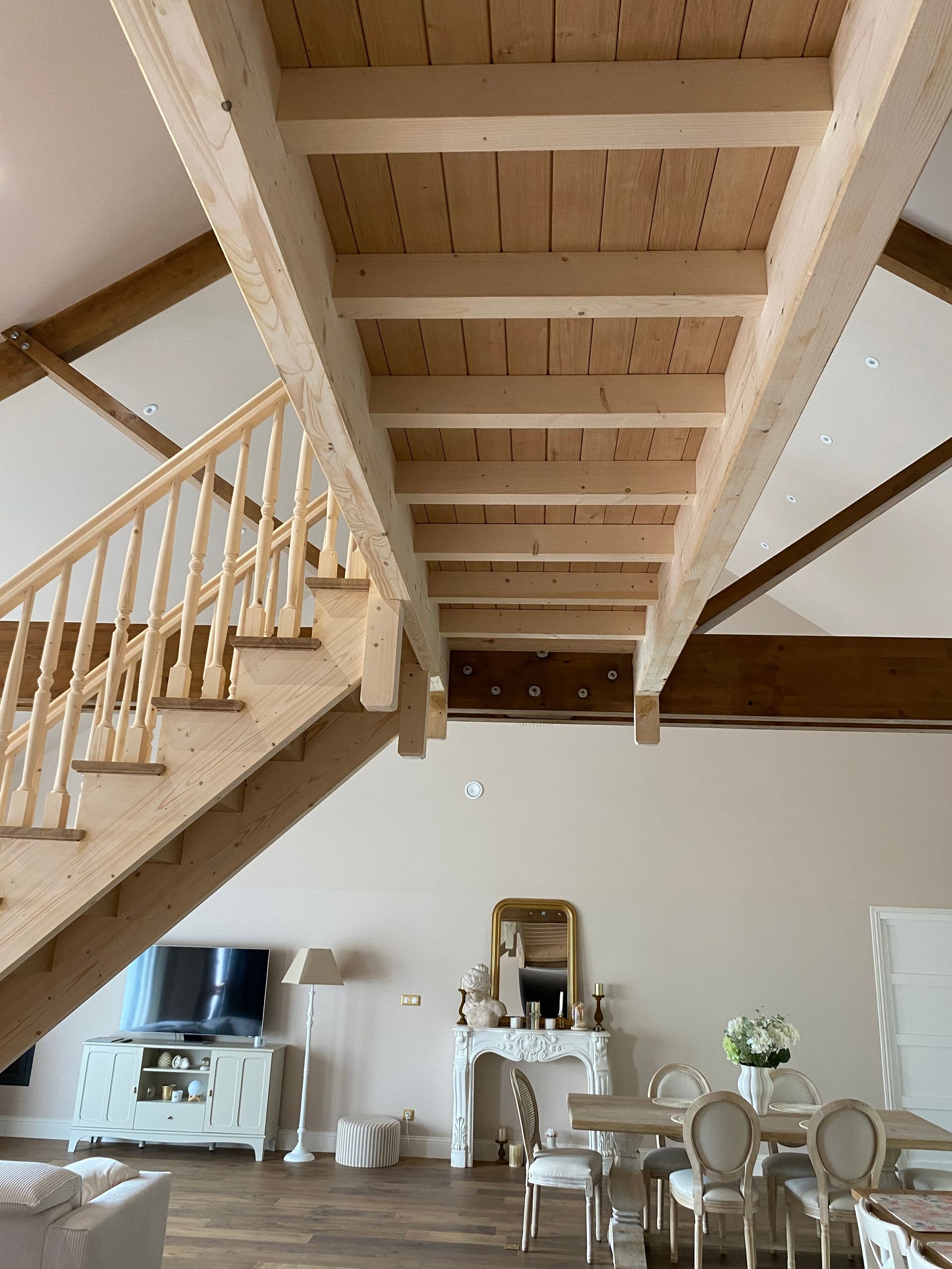 Escalier en bois et poutres apparentes au plafond, au-dessus d’une salle à manger lumineuse avec une table, des chaises et un buffet.