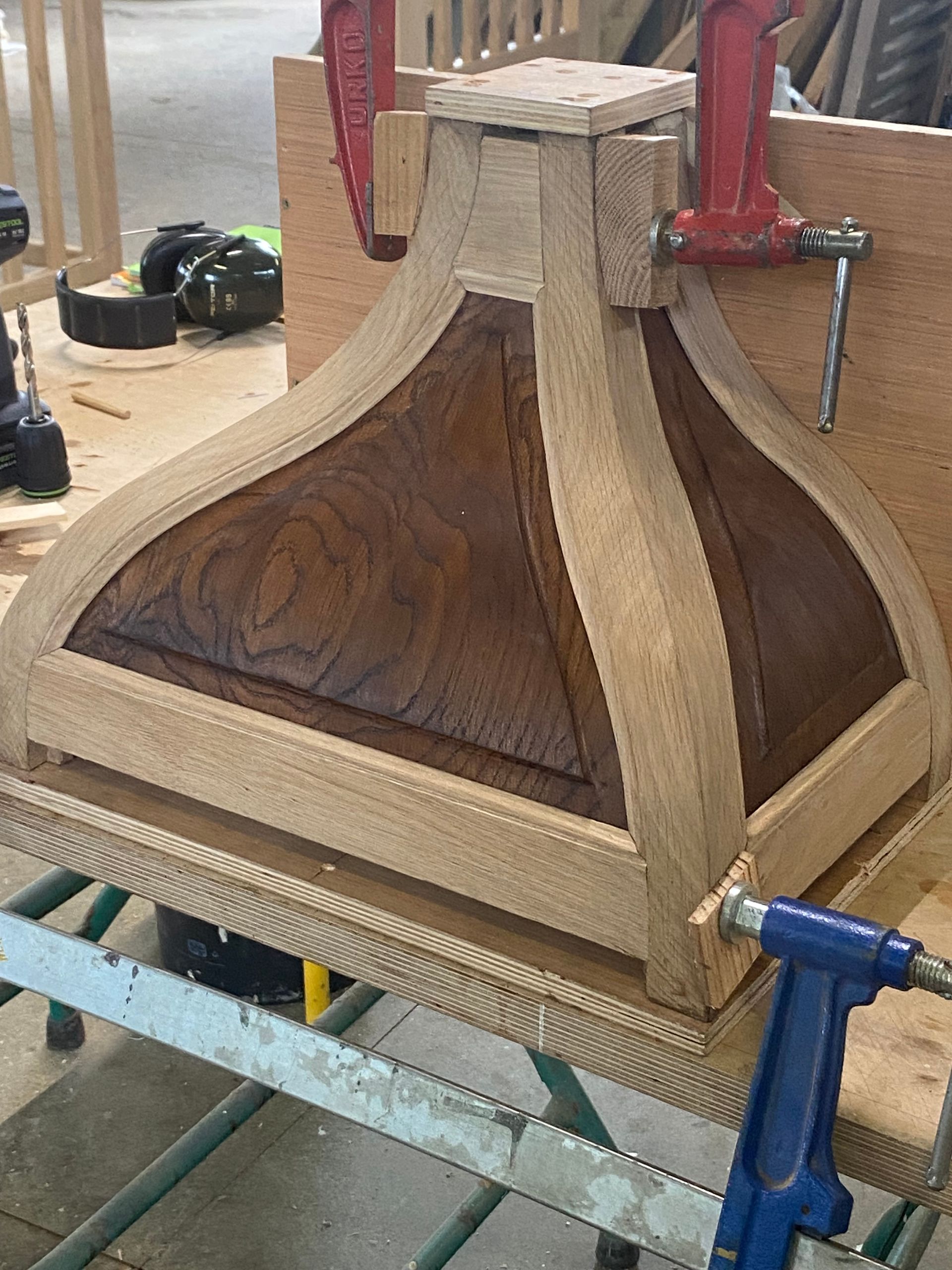 Pièce de mobilier en bois serrée dans un atelier, avec des panneaux incurvés et des serre-joints rouges et bleus fixés dessus.