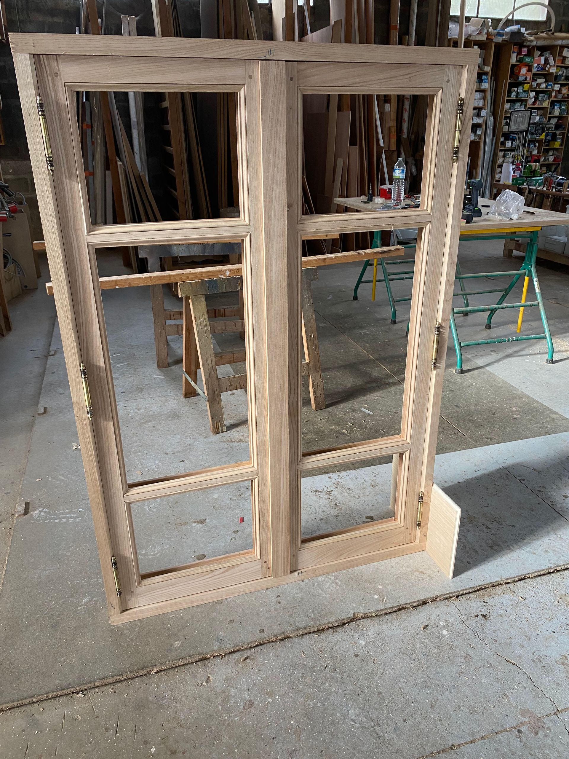 Des cadres de fenêtres en bois empilés dans un atelier, avec des outils et des matériaux visibles en arrière-plan.