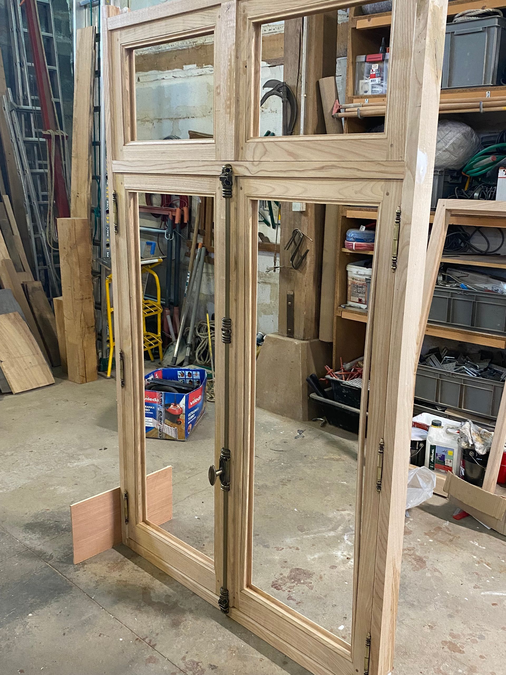 Ensemble de fenêtres et de portes à cadre en bois installé dans un atelier, avec des panneaux de contreplaqué et des outils en arrière-plan.