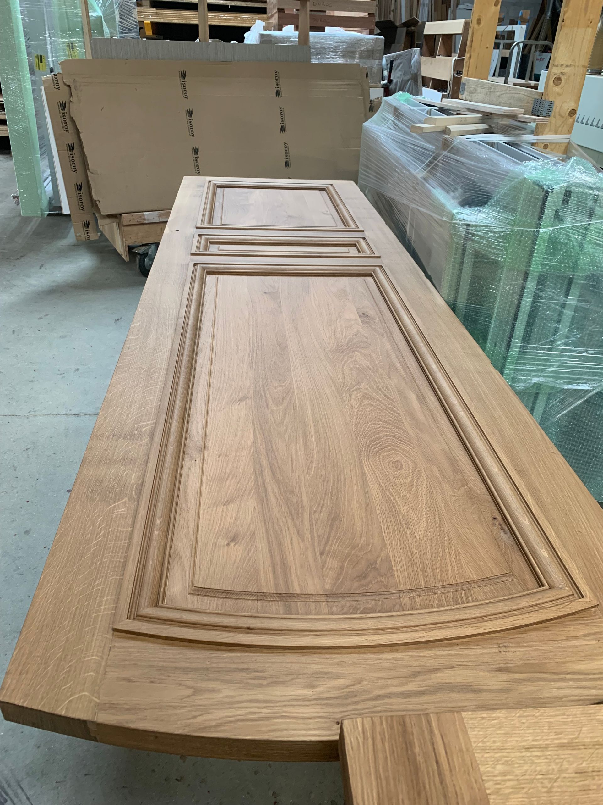 Panneau de porte d’armoire en bois posé sur un établi dans une usine, avec un autre panneau emballé à côté.