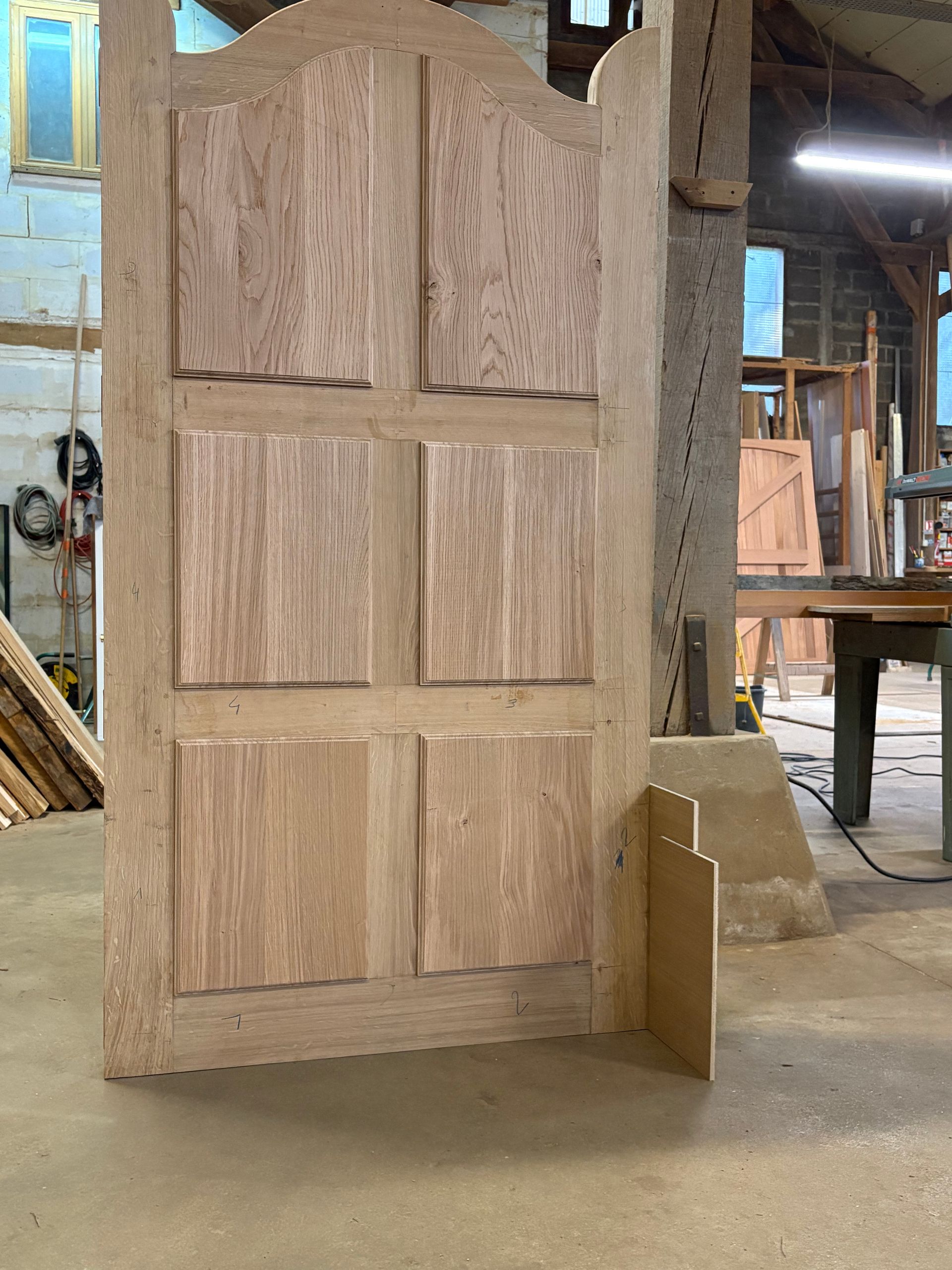 Panneau de porte d’armoire en bois, orné d’arcs sculptés et de sections rectangulaires, appuyé contre un mur dans un atelier.