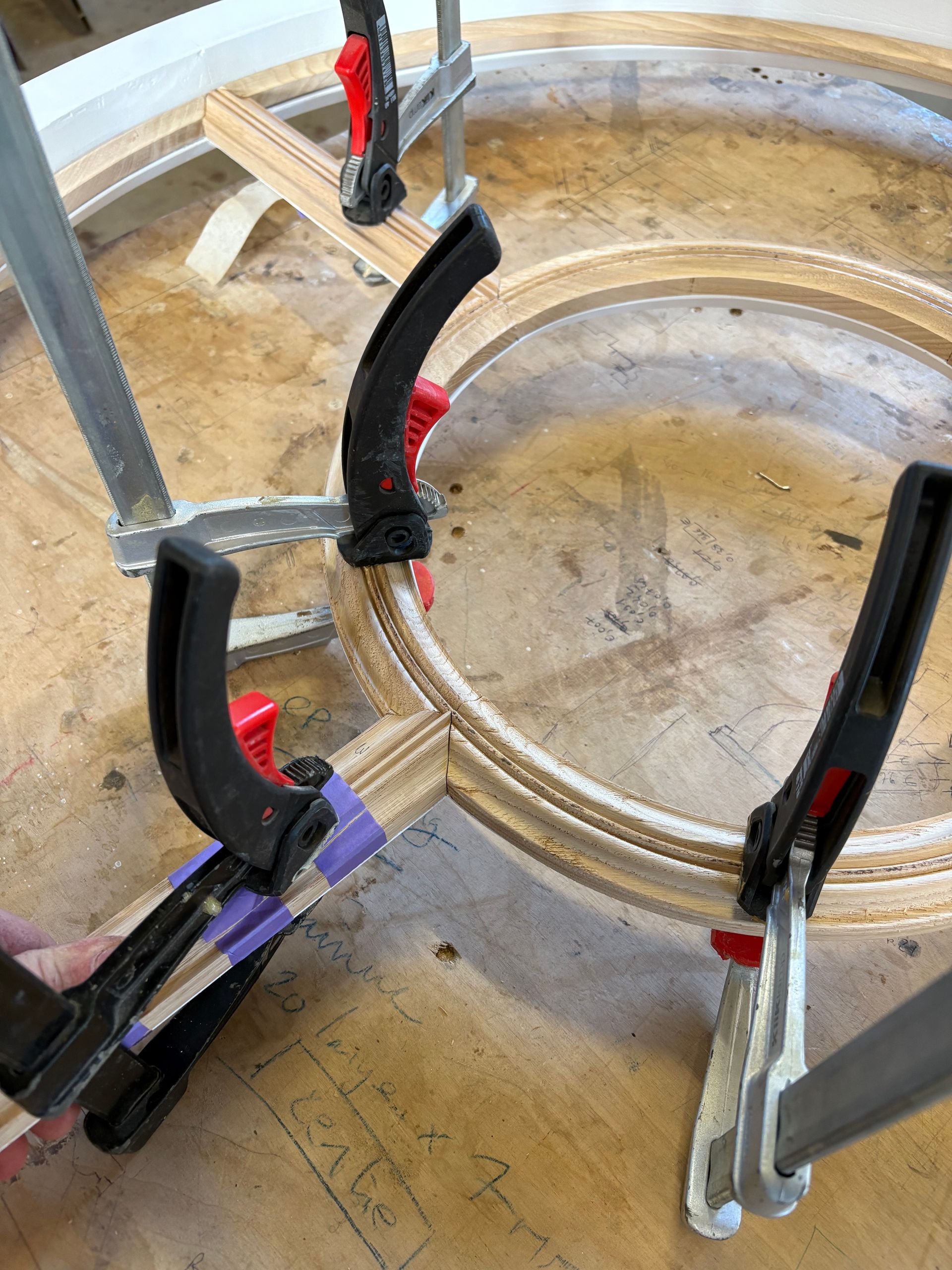 Des serre-joints maintiennent une pièce de bois circulaire sur un établi, avec un outil à manche rouge à proximité.