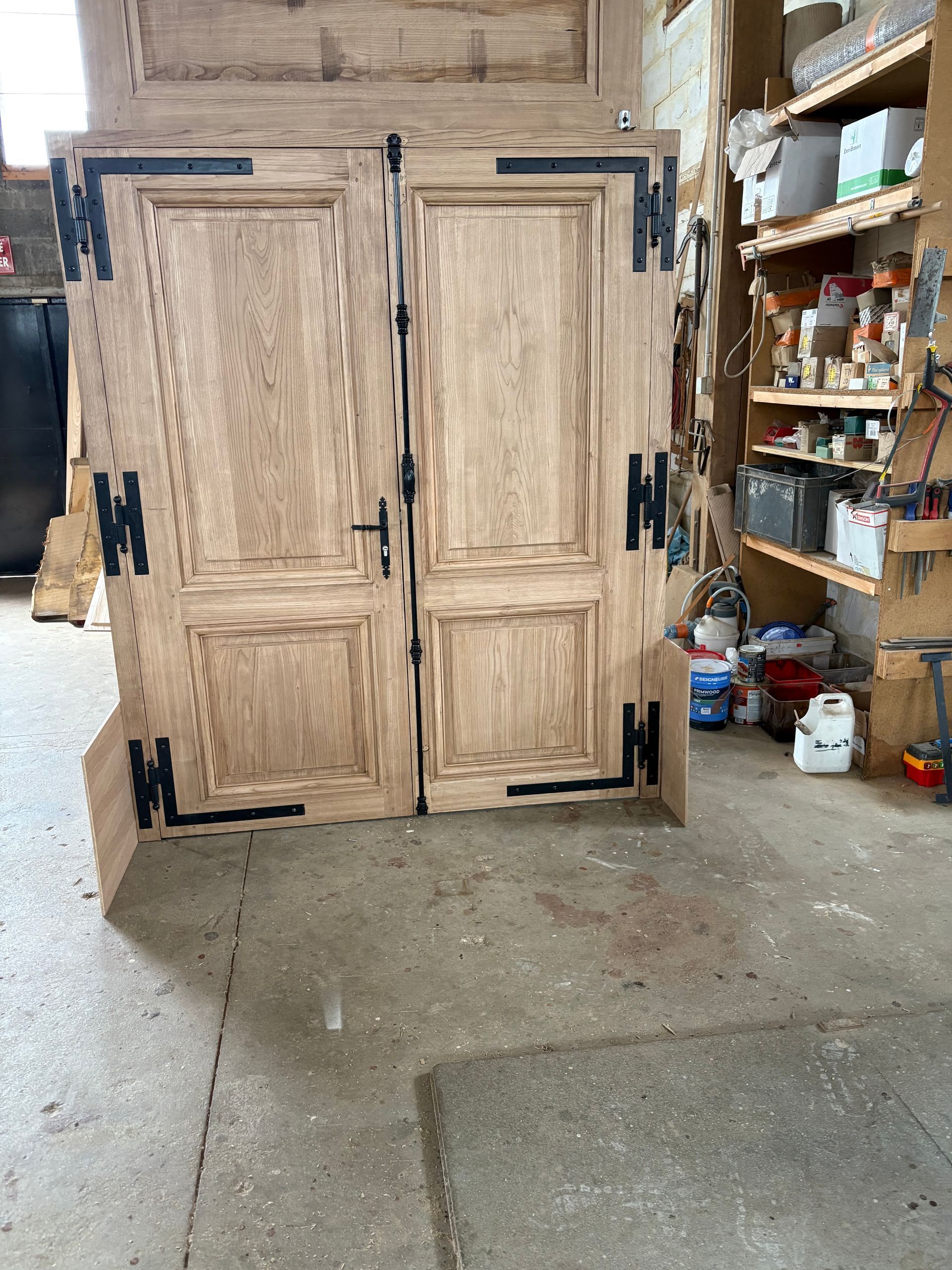 Portes doubles en bois d’un placard dans l’allée d’un atelier, avec des étagères et du matériel en arrière-plan.
