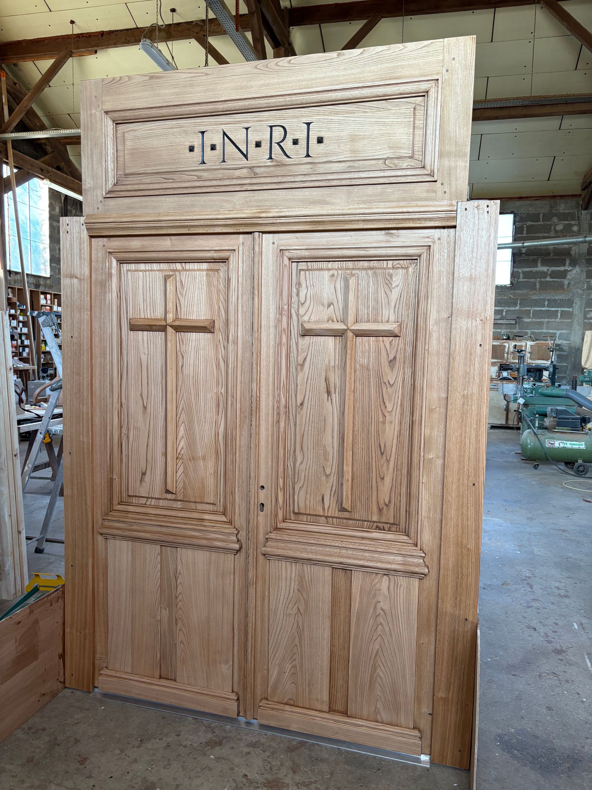 Portes doubles rustiques en bois, ornées de panneaux sculptés, avec l’inscription J.N.R.C. au-dessus, dans un atelier.
