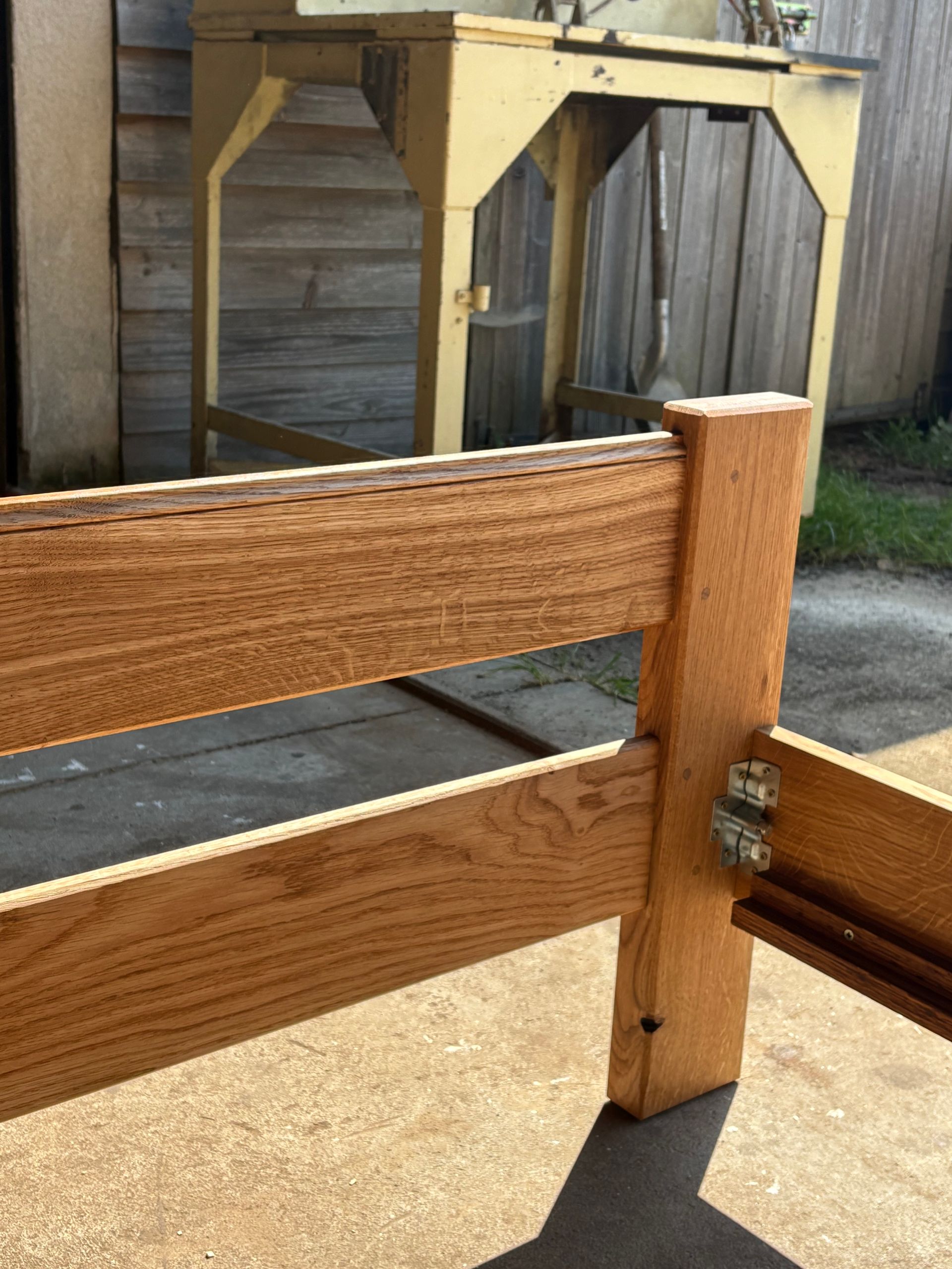 Angle de clôture en bois avec un loquet à charnière métallique, à côté d’une terrasse en béton et d’une enceinte grillagée.