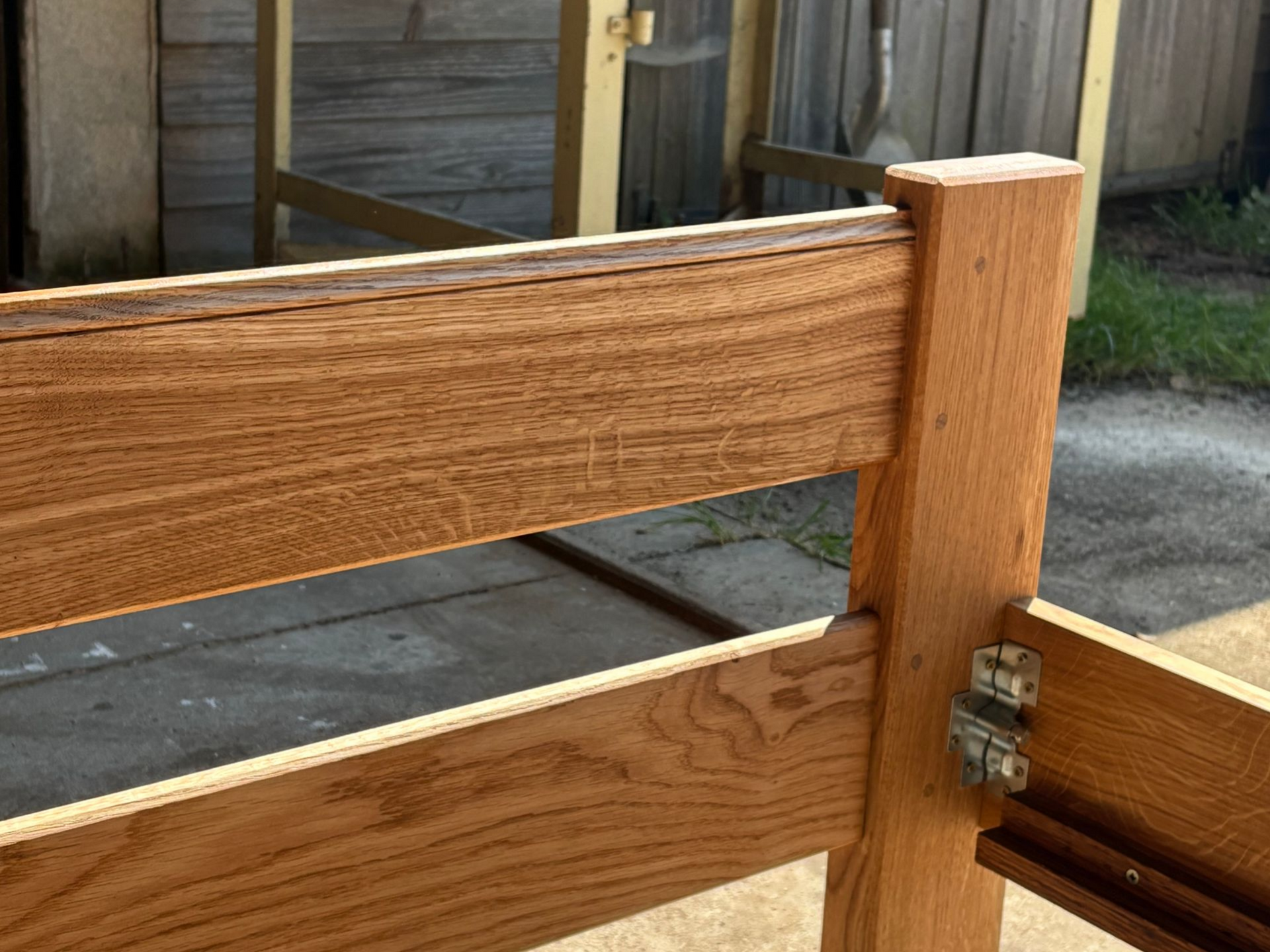 Lit en bois à lames horizontales, avec une charnière métallique, près d’une terrasse clôturée.