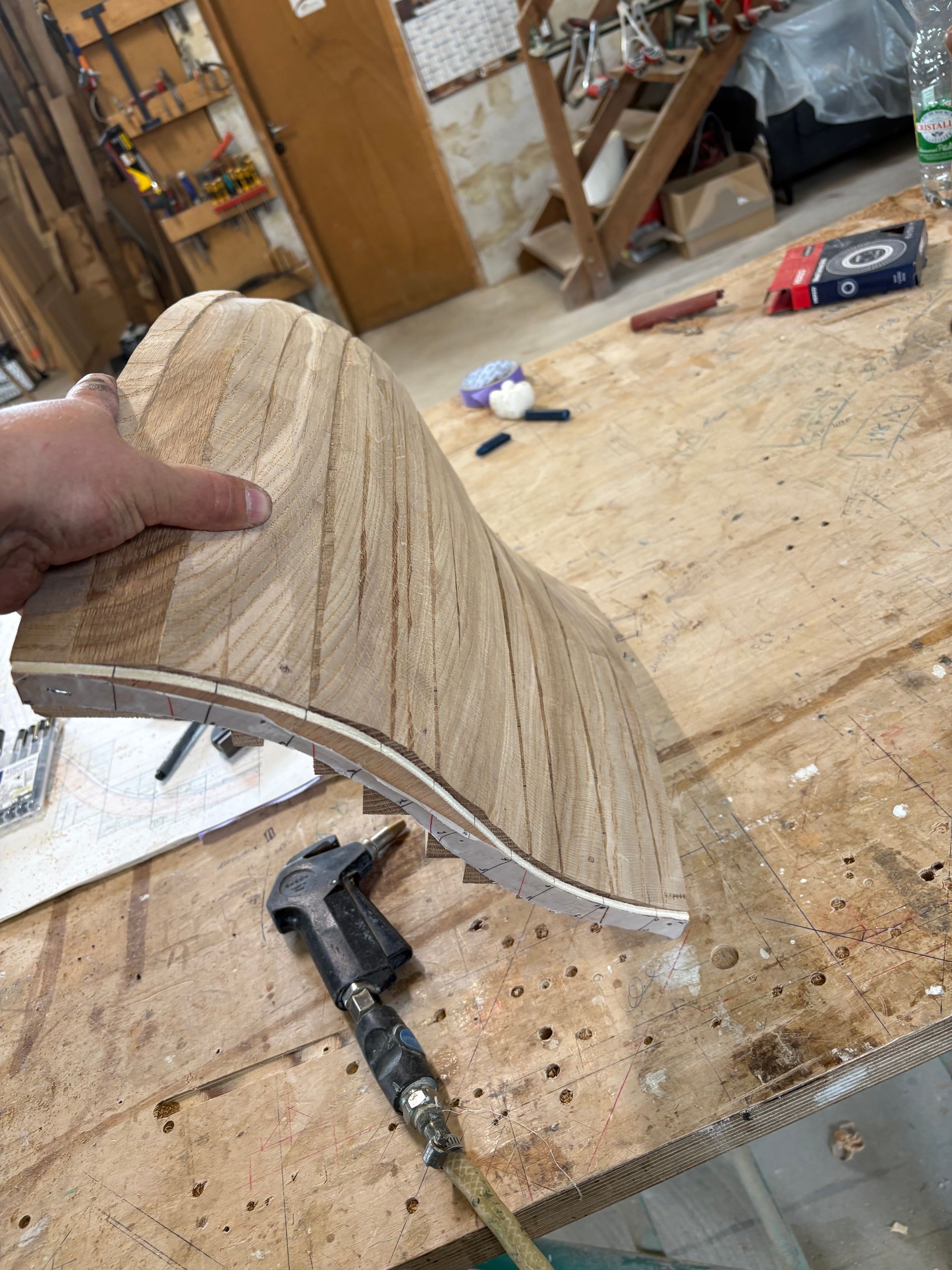 Une main tient une pièce de bois stratifié incurvée au-dessus d’un établi, dans un atelier.