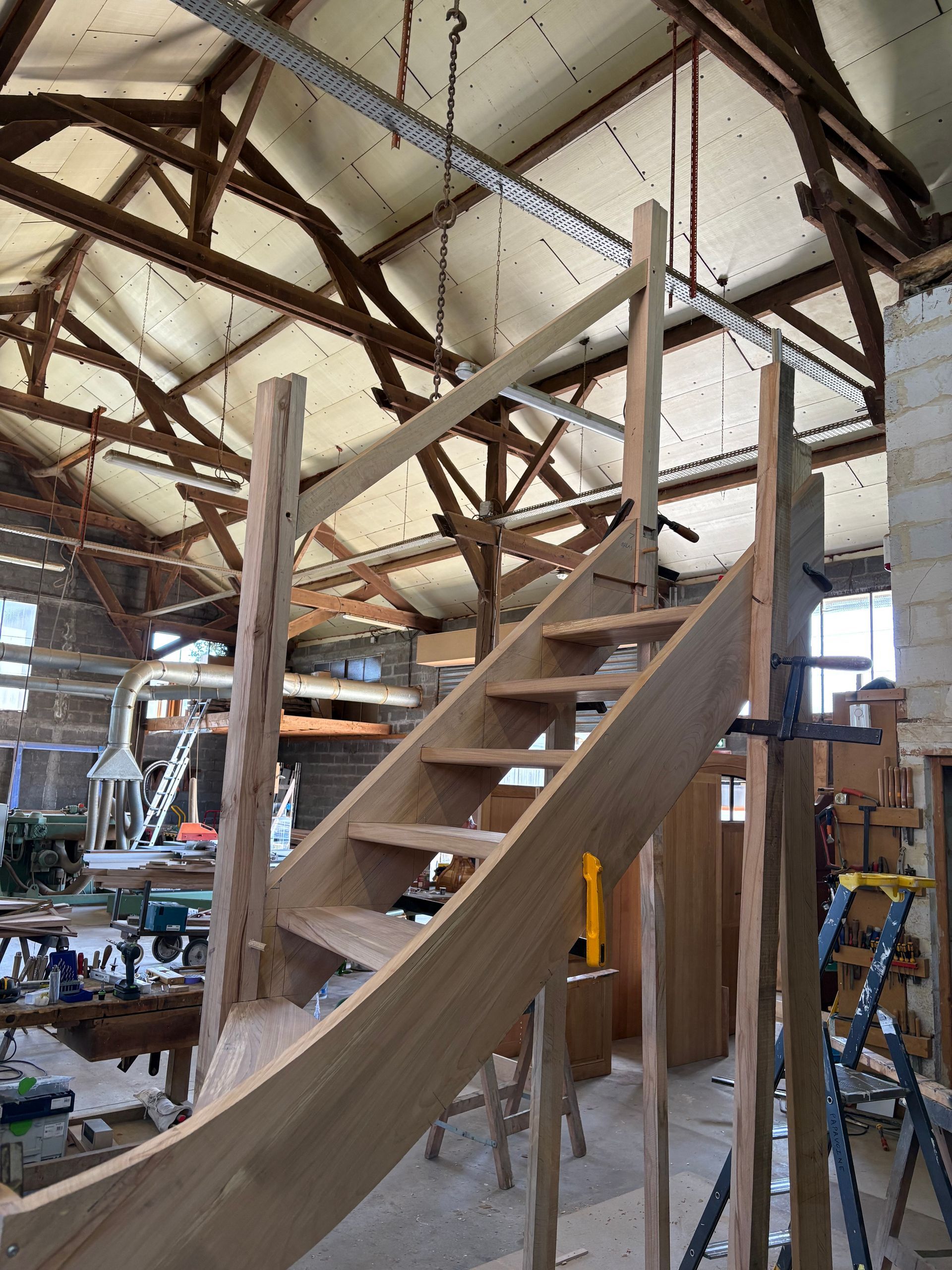 Escalier en bois dans un atelier rustique avec des poutres apparentes et des outils tout autour.