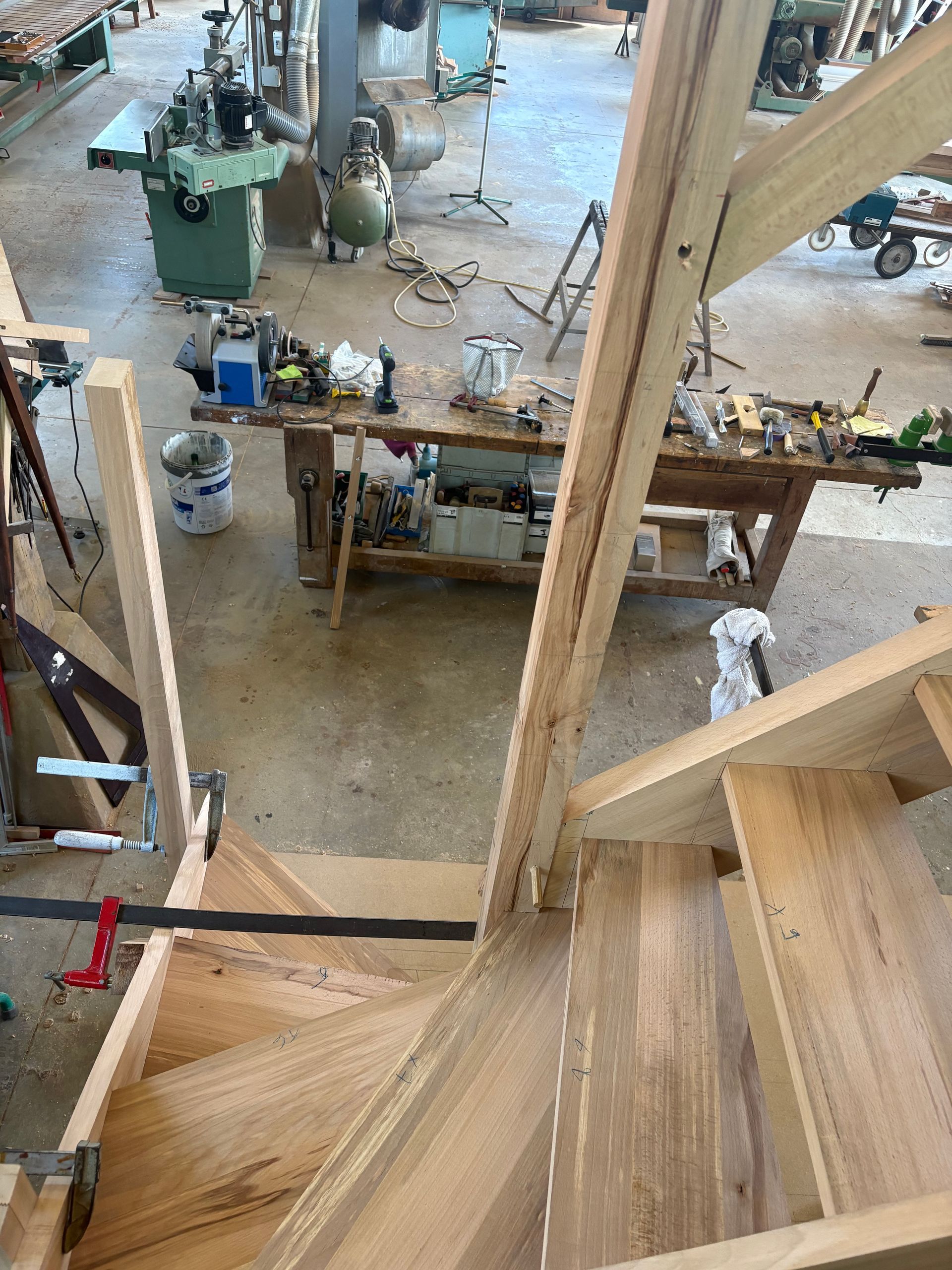 Escalier en bois surplombant un atelier animé, avec des machines et des outils au rez-de-chaussée.