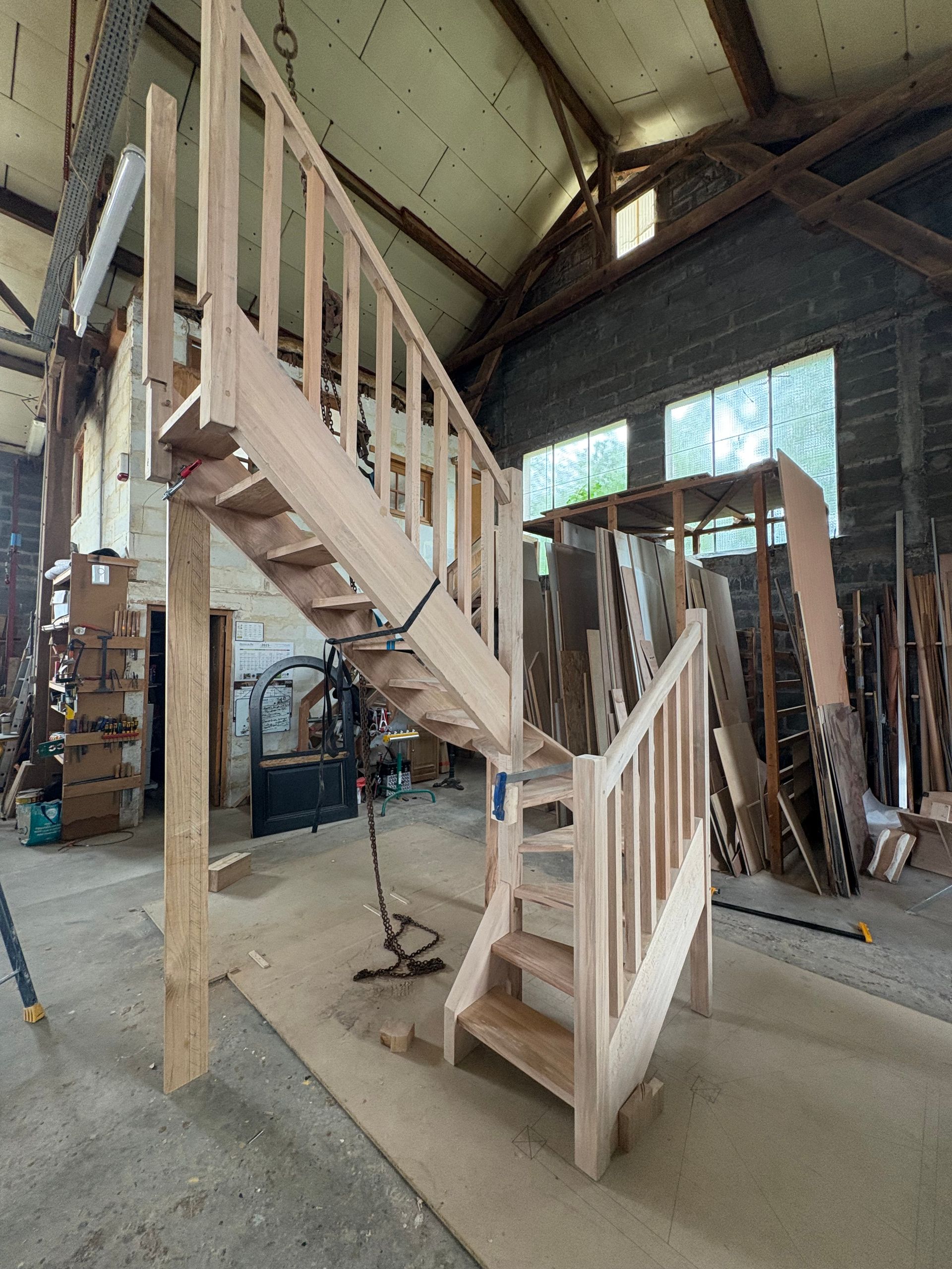 Escalier en bois partiellement construit à l’intérieur d’un grand atelier avec des poutres apparentes et des échelles.