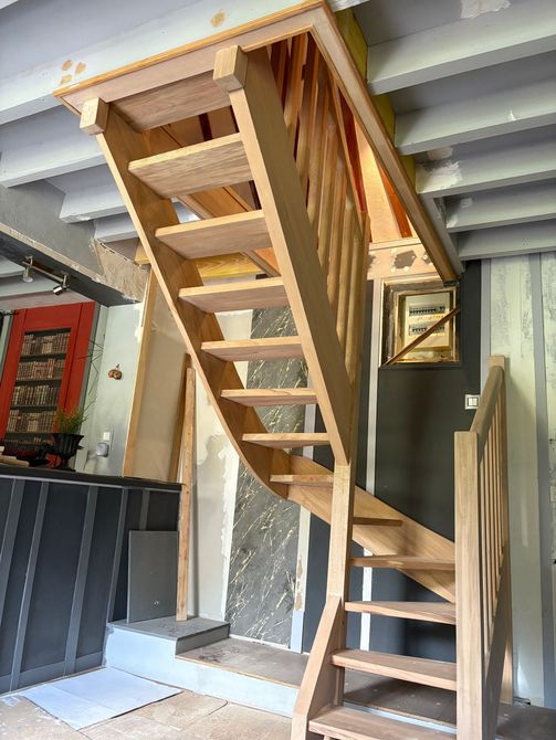 Escalier raide en bois menant aux combles, avec des marches ouvertes et une rampe, dans un espace intérieur étroit.