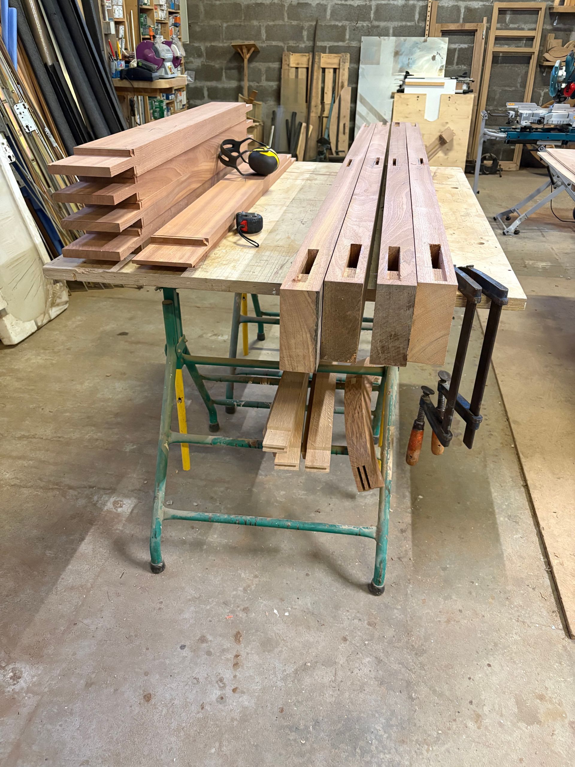 Du bois de construction empilé et de longues poutres en bois sur un établi vert dans un atelier de menuiserie.