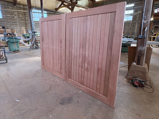 Grand panneau de portail en bois brut inachevé dans un atelier, avec des outils et du bois de construction en arrière-plan.