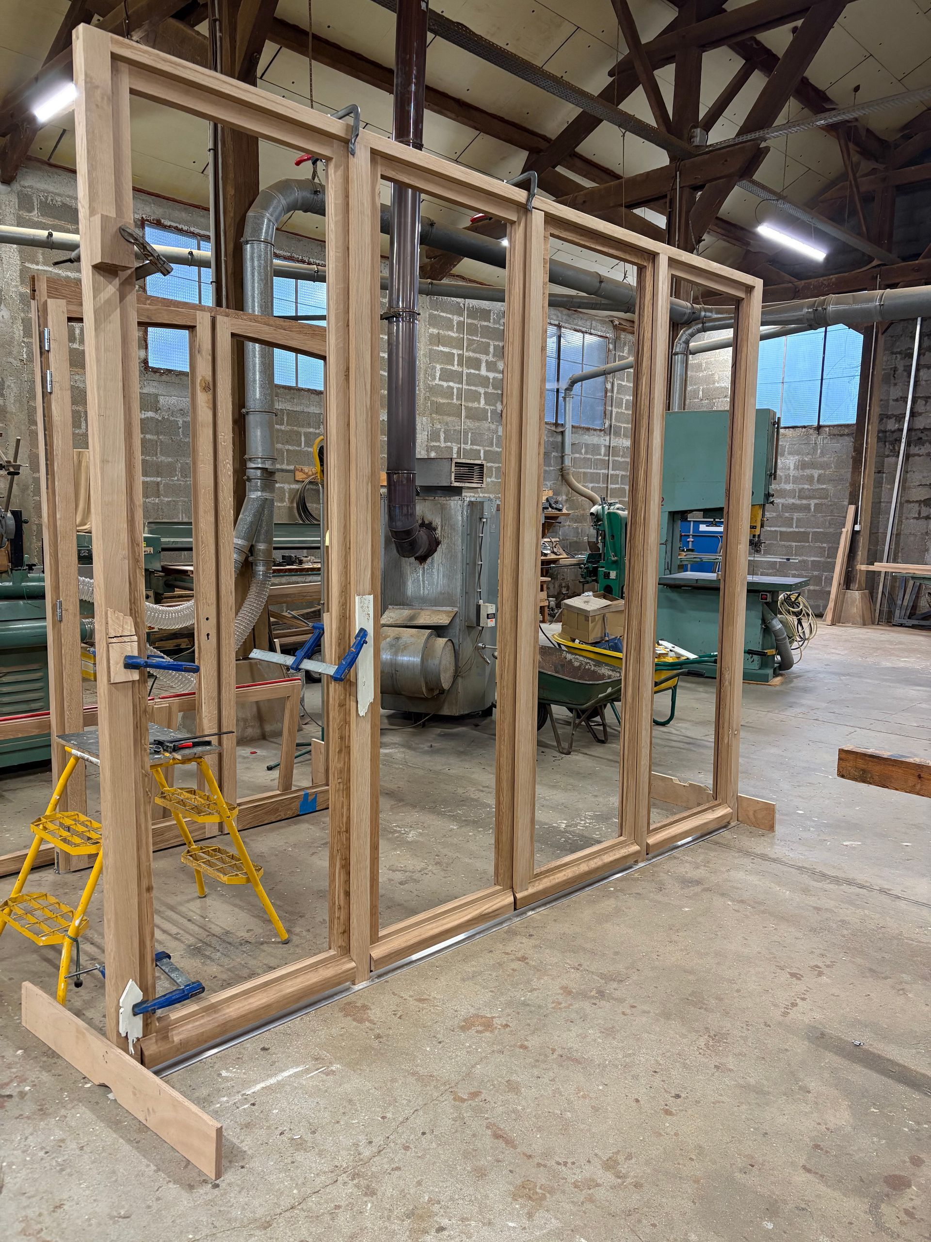 Cadre d'une baie vitrée en bois en cours de construction dans un atelier, avec des outils et du bois de construction en arrière-plan.