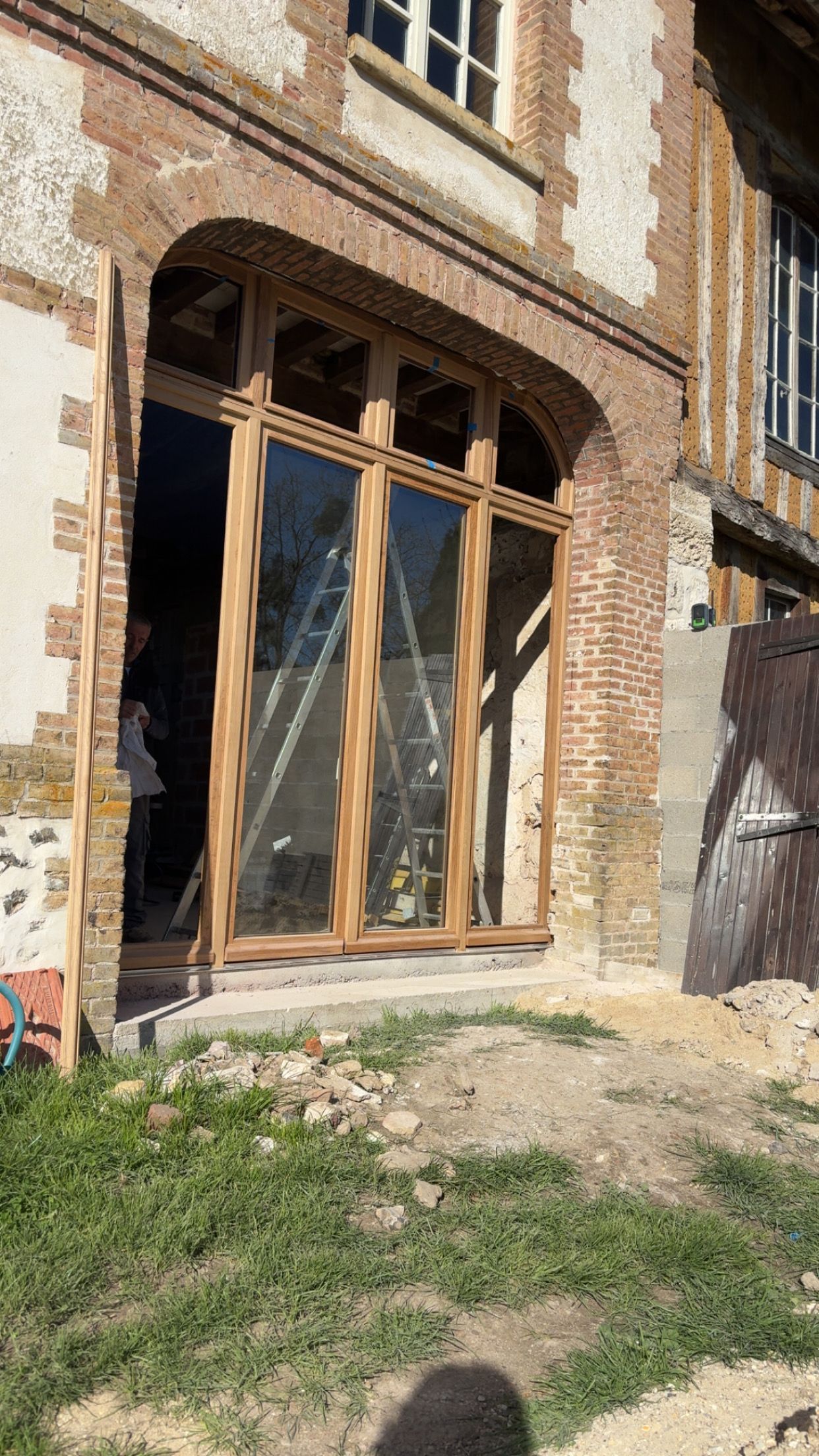 Façade d’un ancien bâtiment en brique avec une grande porte encadrée de bois en cours de construction, recouverte d’une bâche en plastique.