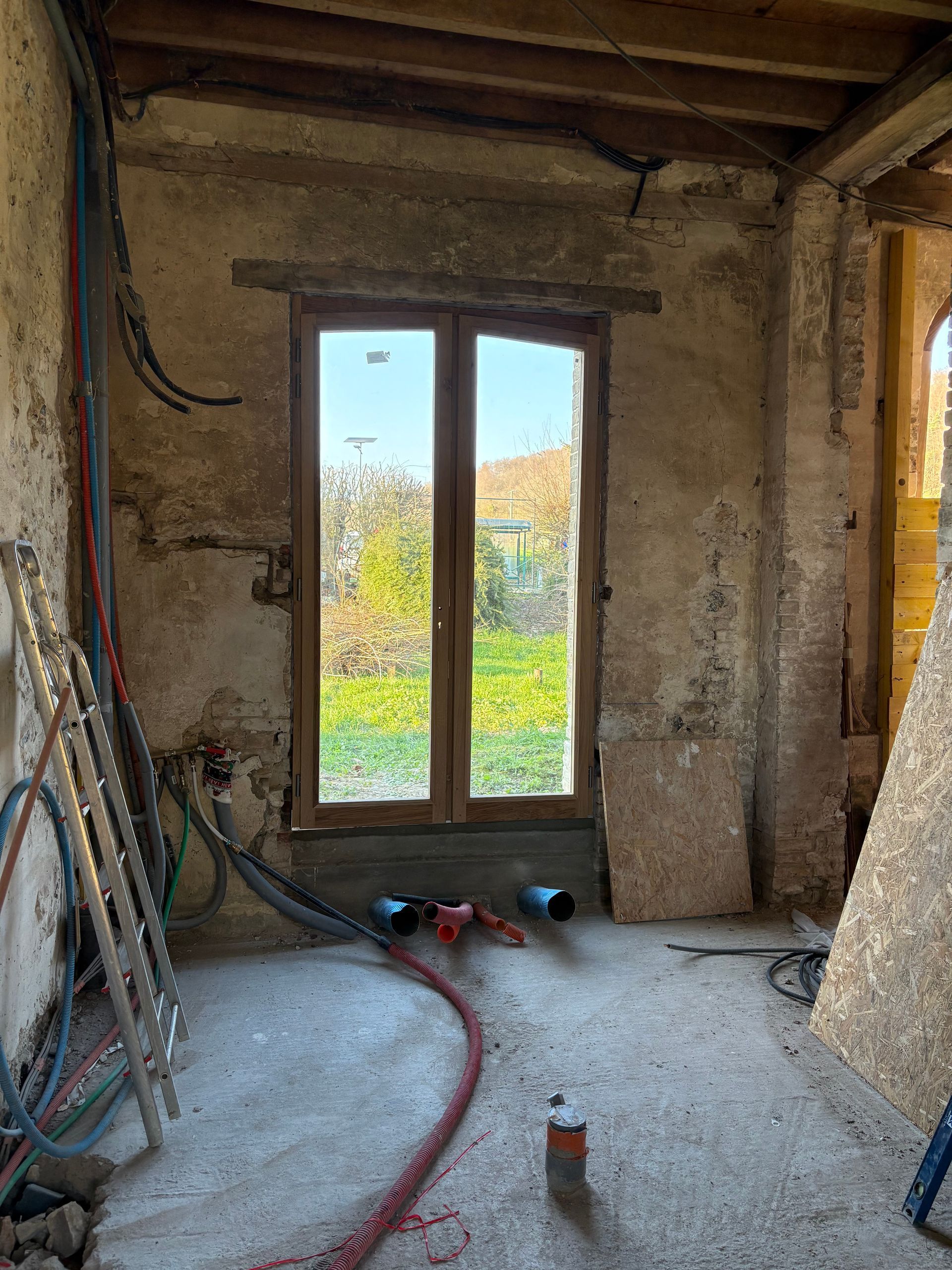 Pièce poussiéreuse en travaux, avec une fenêtre, des outils au sol et des fils électriques apparents sur le mur de gauche.