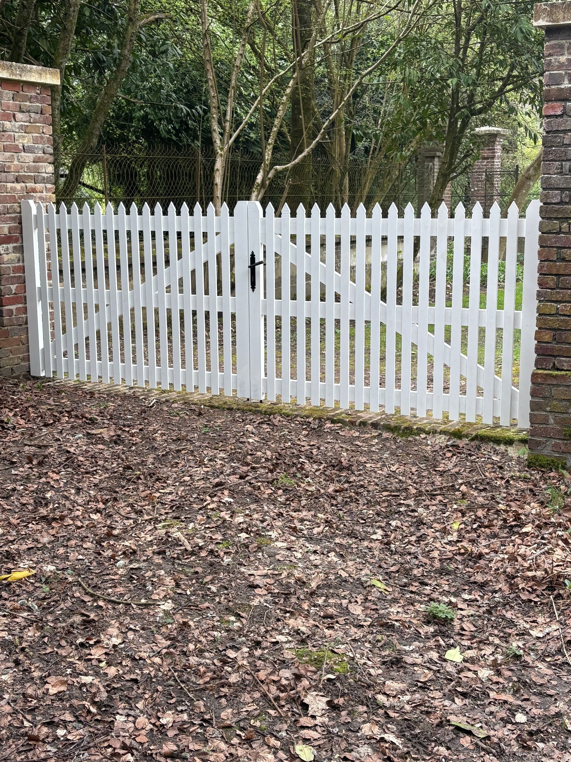 Une clôture blanche à piquets avec un portail dans un jardin boisé jonché de feuilles mortes
