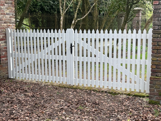 Portail blanc à piquets, renforcé par des traverses diagonales et encadré de piliers en brique, au bord d’un sentier boisé.