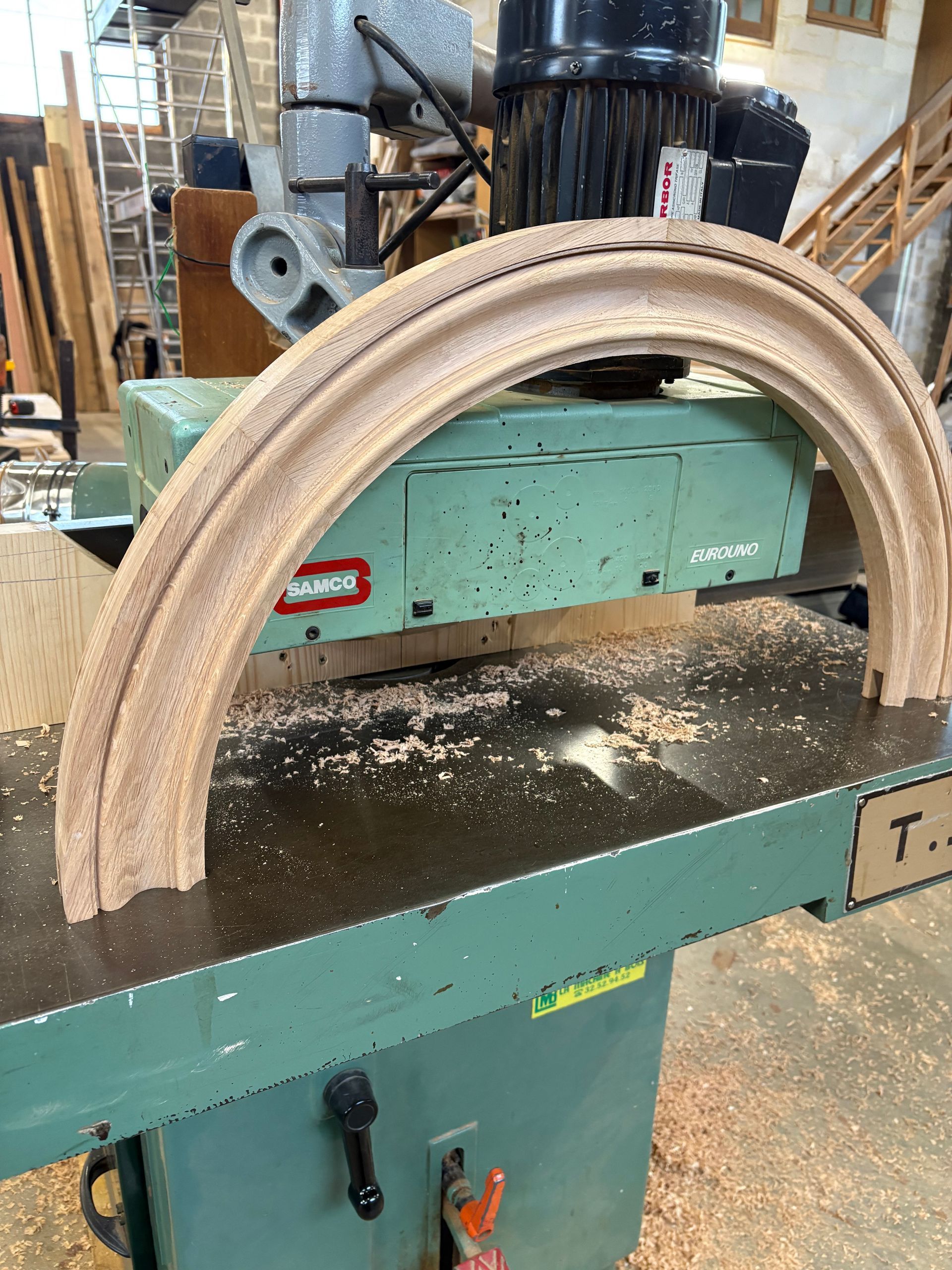 Atelier de menuiserie avec une planche courbée est découpée sur une grande machine, avec de la sciure éparpillée en dessous.