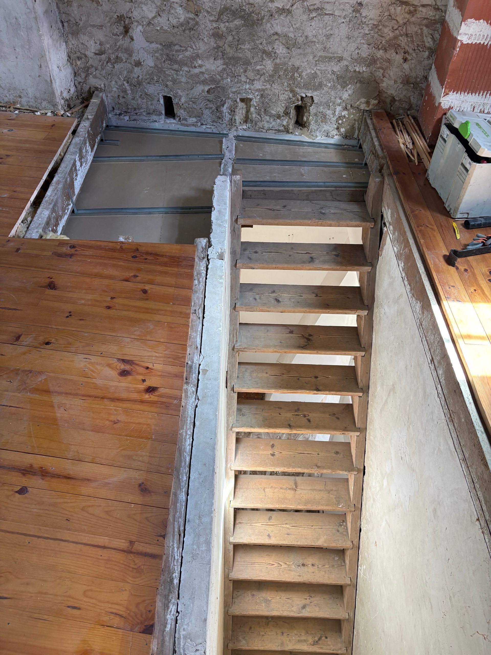 Vue plongeante sur une cage d’escalier inachevée, avec des marches en bois apparentes et des murs intérieurs usés.