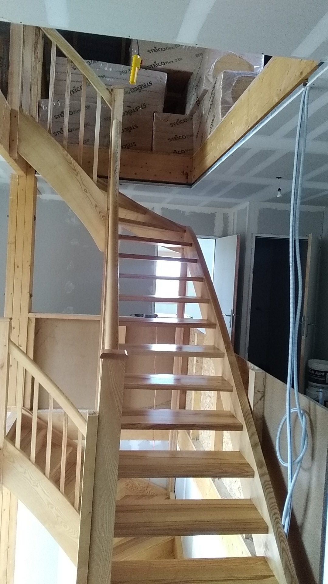 Escalier en bois menant à un étage supérieur en mezzanine, avec une rambarde et des cartons, dans un intérieur non aménagé.