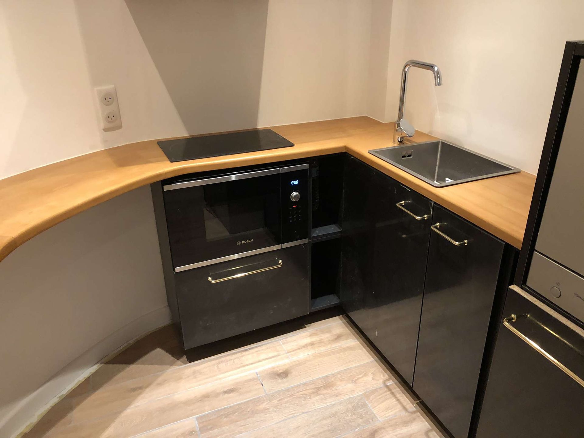 Cuisinette avec plan de travail en bois, placards noirs, évier, micro-ondes et réfrigérateur dans une petite pièce.