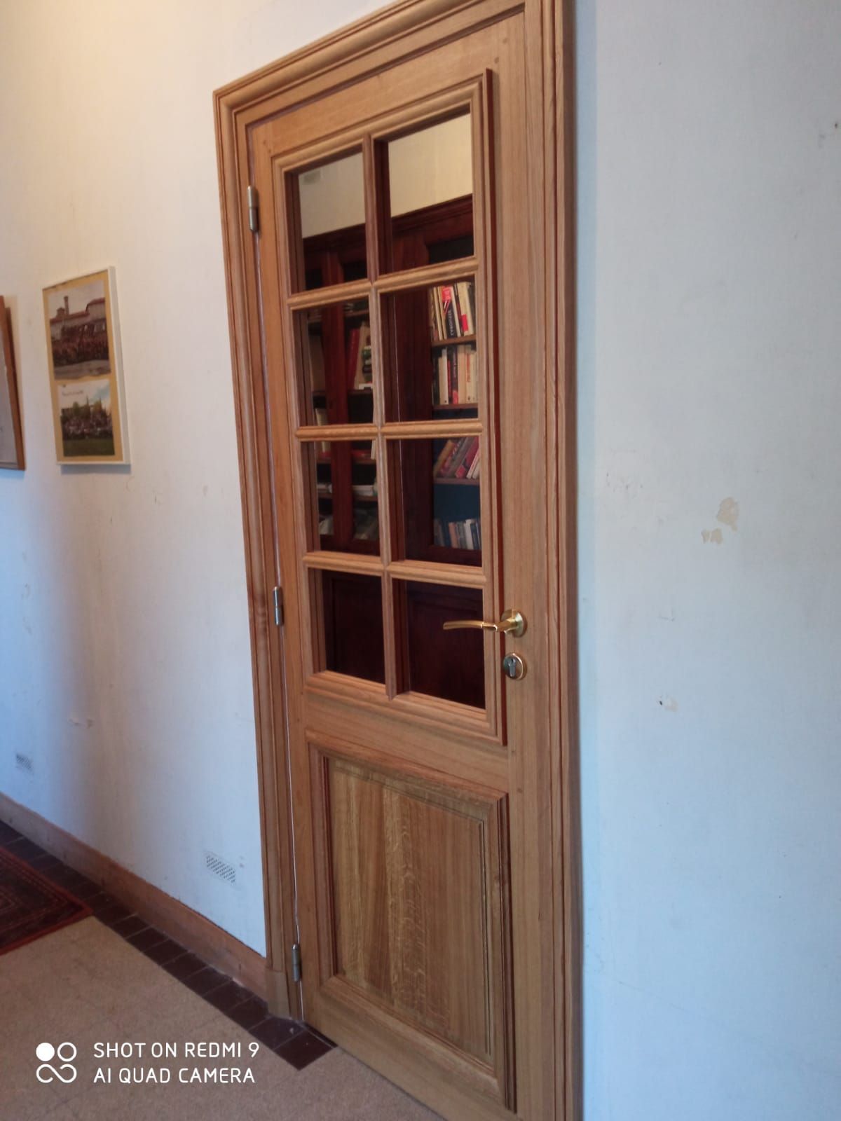 Porte en bois vitrée dans un couloir aux murs blancs, ornés d’œuvres encadrées.