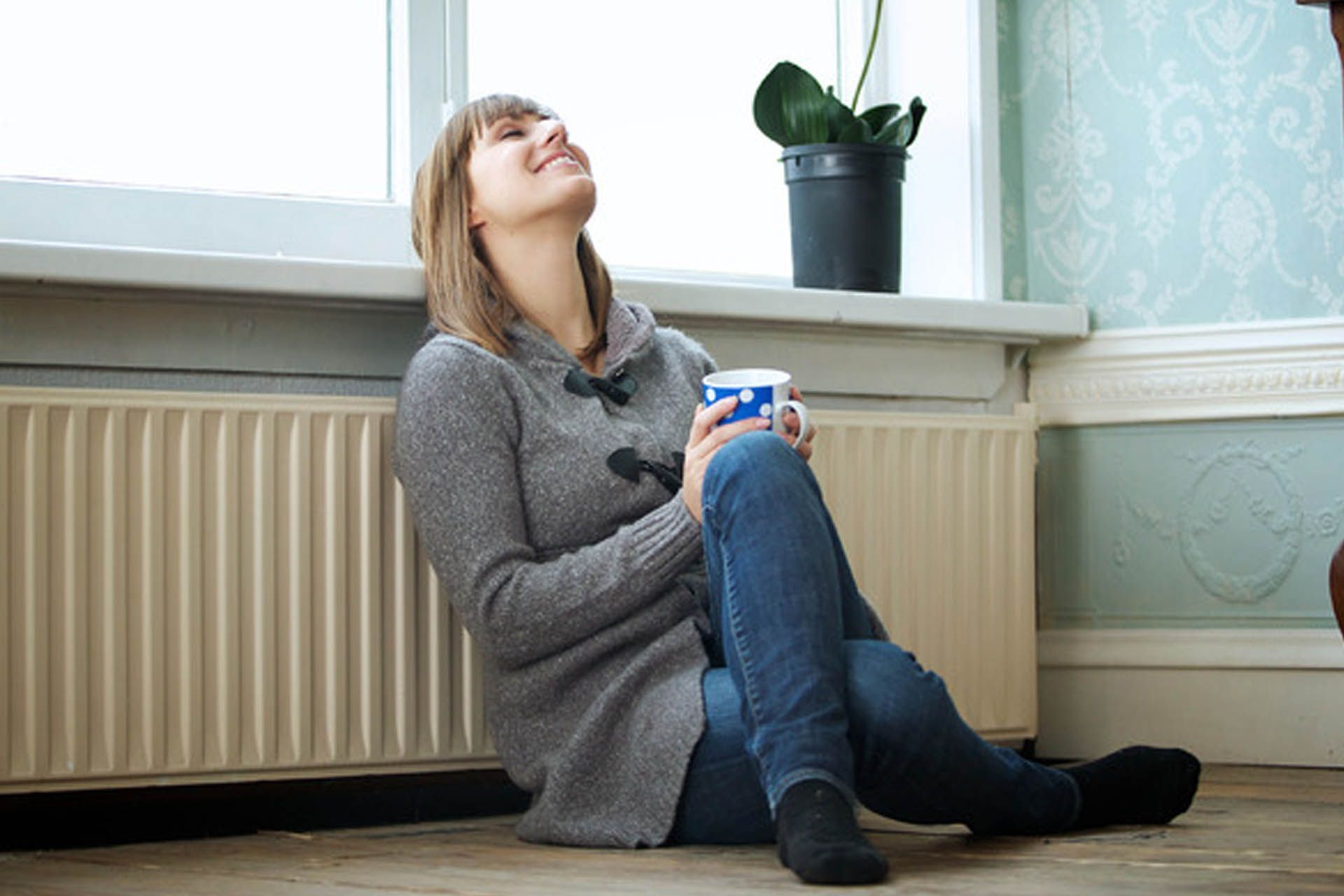 Una mujer está sentada en el suelo junto a un radiador sosteniendo una taza de café.