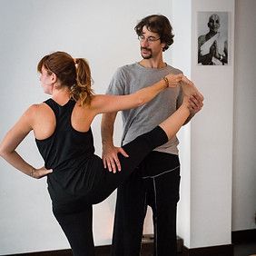 Mujer en pose de yoga, instructor ayudando con el estiramiento de piernas, habitación llena de luz.