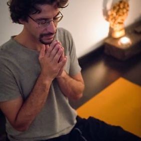 Hombre con gafas, manos juntas, mirando hacia abajo. Interior con vela y esterilla de yoga.
