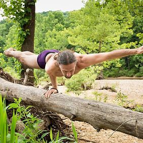 Un hombre con pantalones cortos de color morado hace una pose de yoga sobre un tronco caído en un bosque.