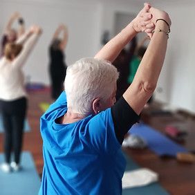 Mujer con camisa azul dirigiendo una clase de yoga, con los brazos levantados e inclinados hacia arriba