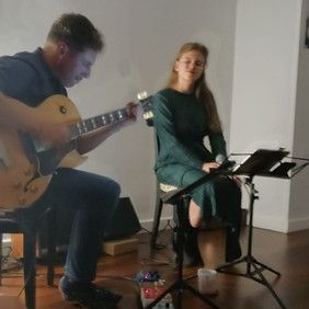 Un hombre toca la guitarra mientras una mujer canta con un micrófono. Están en un espacio cerrado, interpretando música.