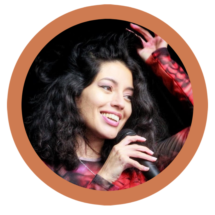 Una mujer con cabello oscuro y rizado sonríe mientras sostiene un micrófono; lleva una blusa estampada.