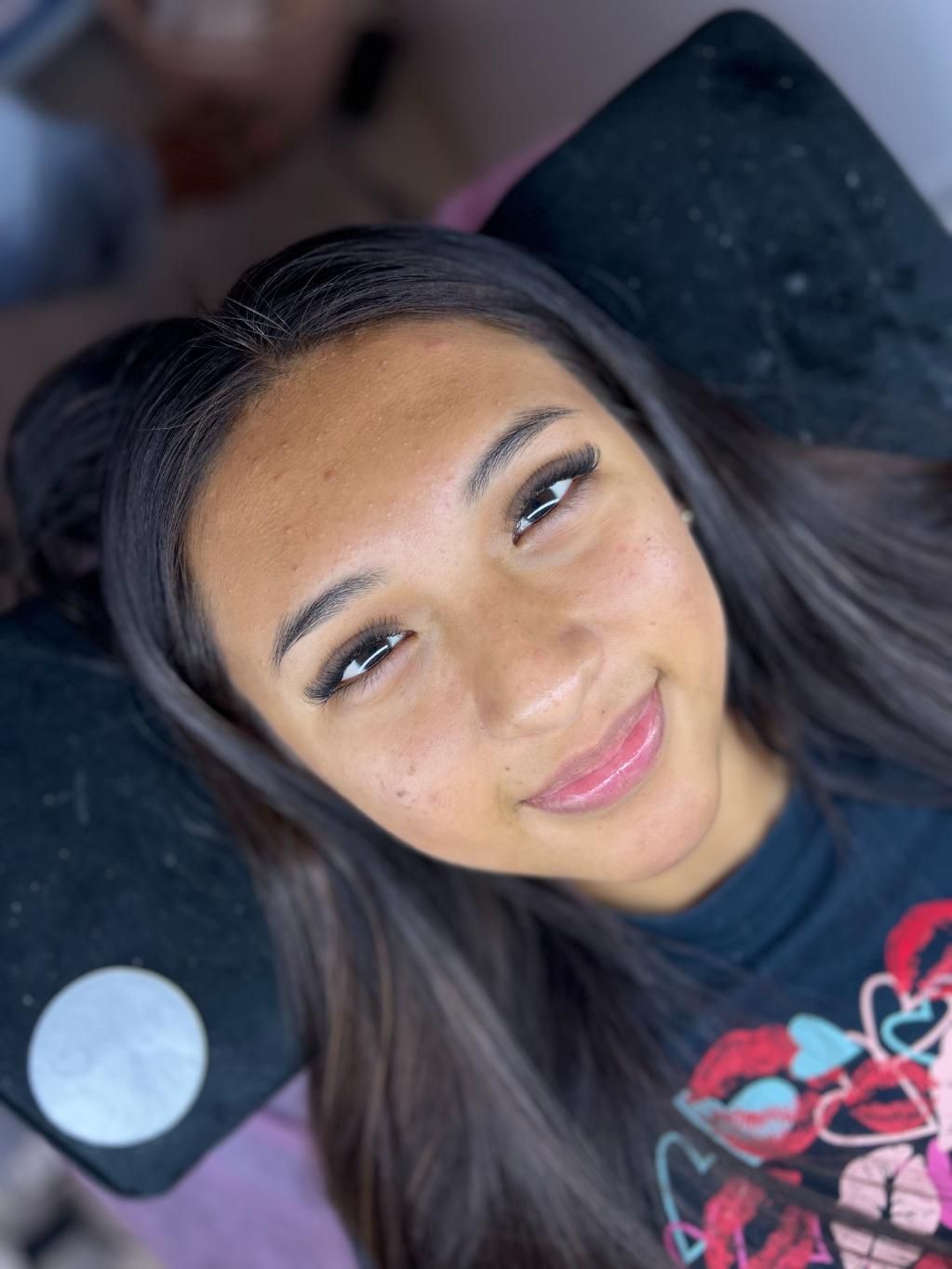 Mujer con cabello oscuro y extensiones de pestañas sonríe, acostada sobre una almohada negra.