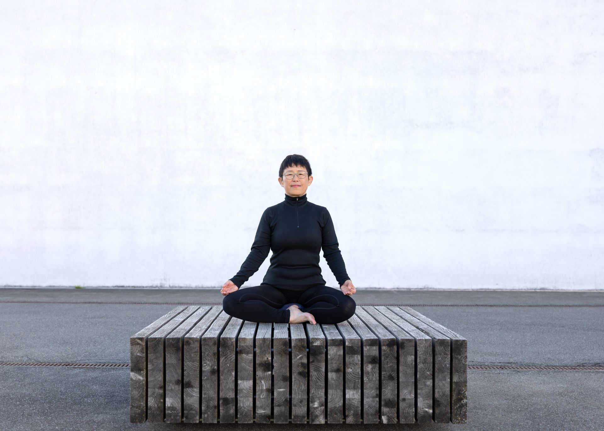 Frau in schwarzem Outfit meditiert auf einer Holzplattform vor einer weißen Wand.