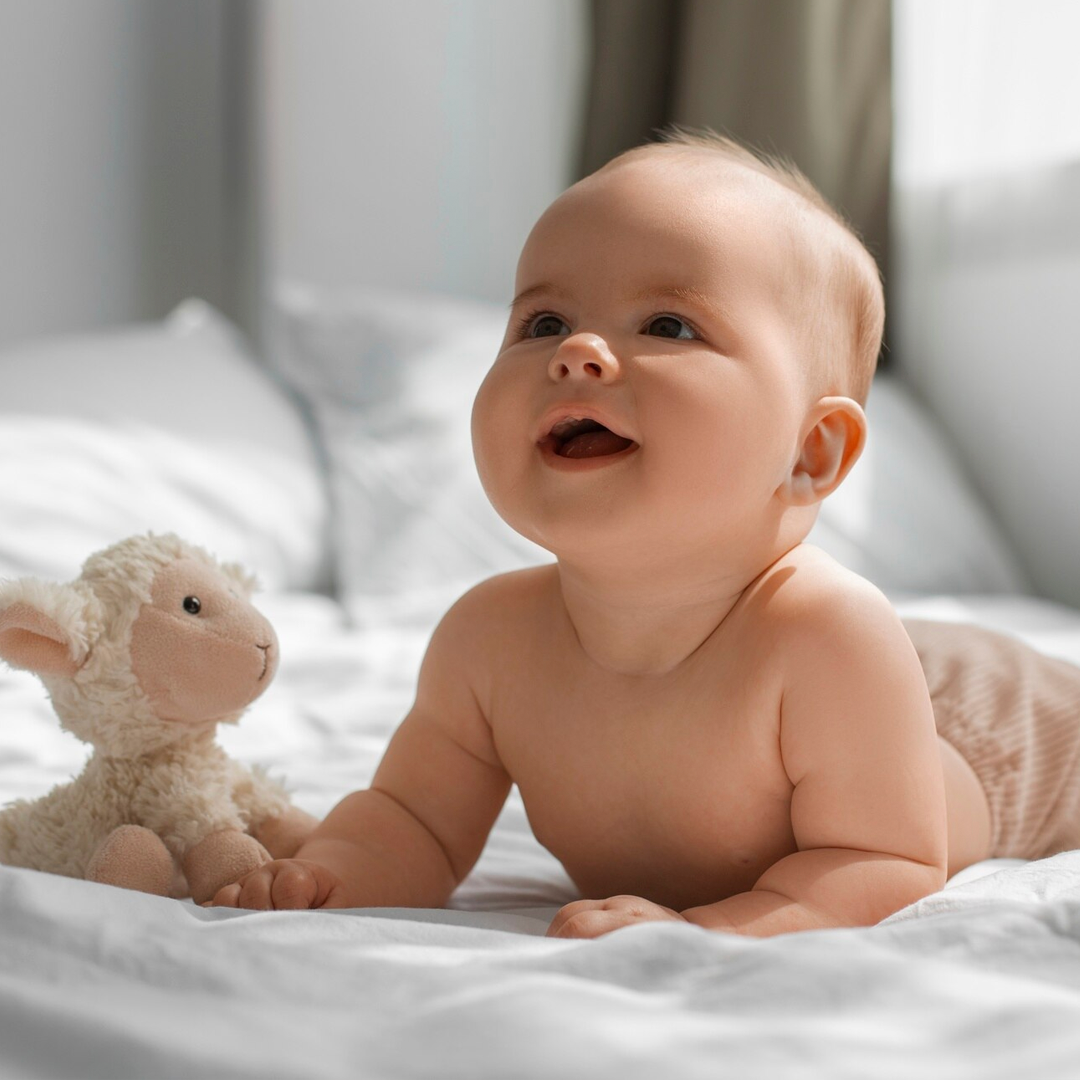 Un bambino a pancia in giù sorride a qualcosa fuori dallo schermo, un agnellino giocattolo accanto, su una coperta bianca.